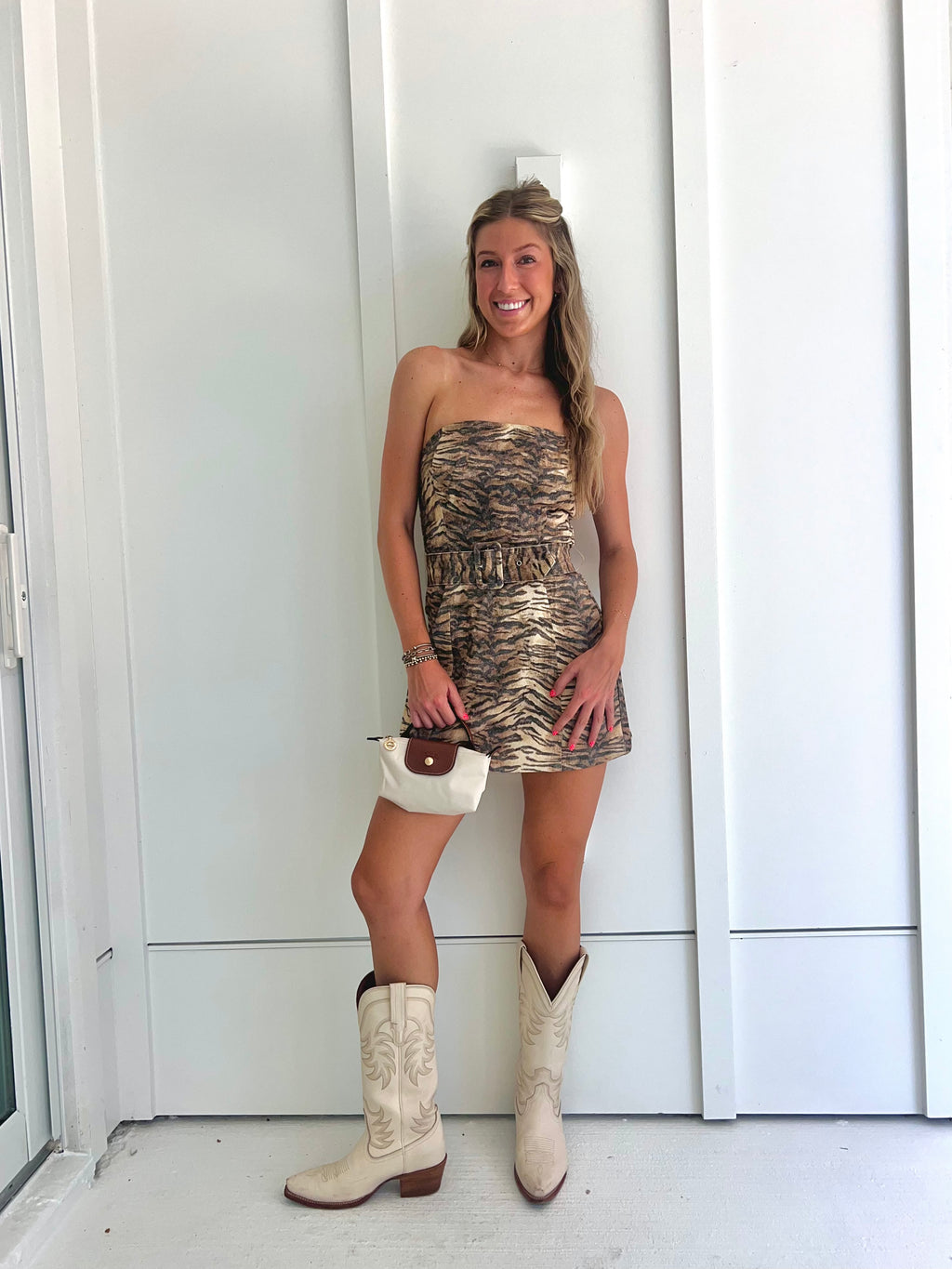 A woman wearing a strapless tiger printed dress and cream cowgirl boots with a cream purse standing against a white wall.