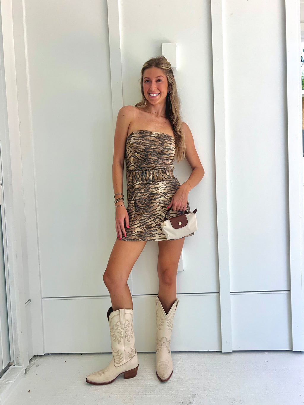 A woman wearing a strapless tiger printed dress and cream cowgirl boots with a cream purse standing against a white wall.