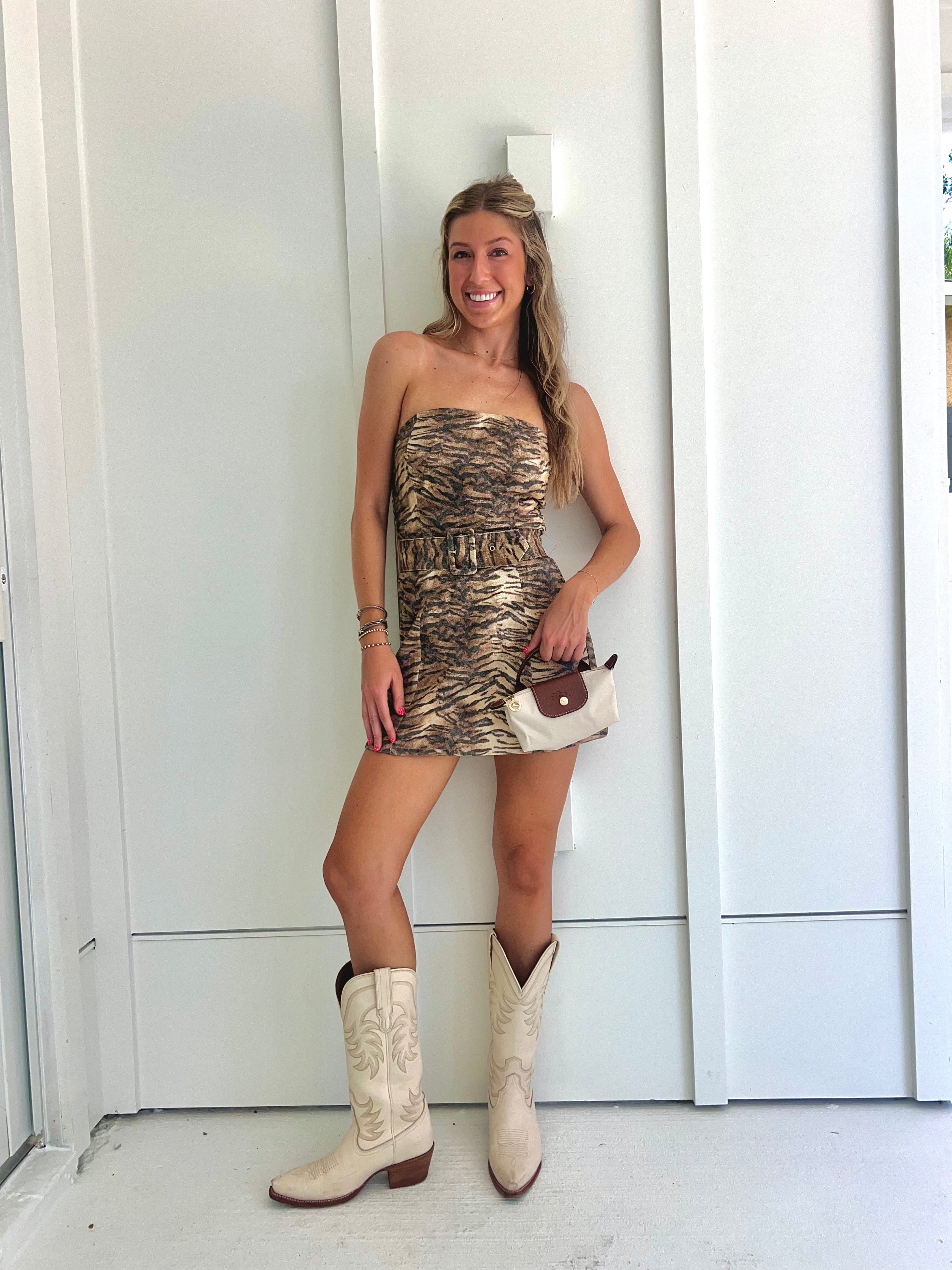 A woman wearing a strapless tiger printed dress and cream cowgirl boots with a cream purse standing against a white wall.