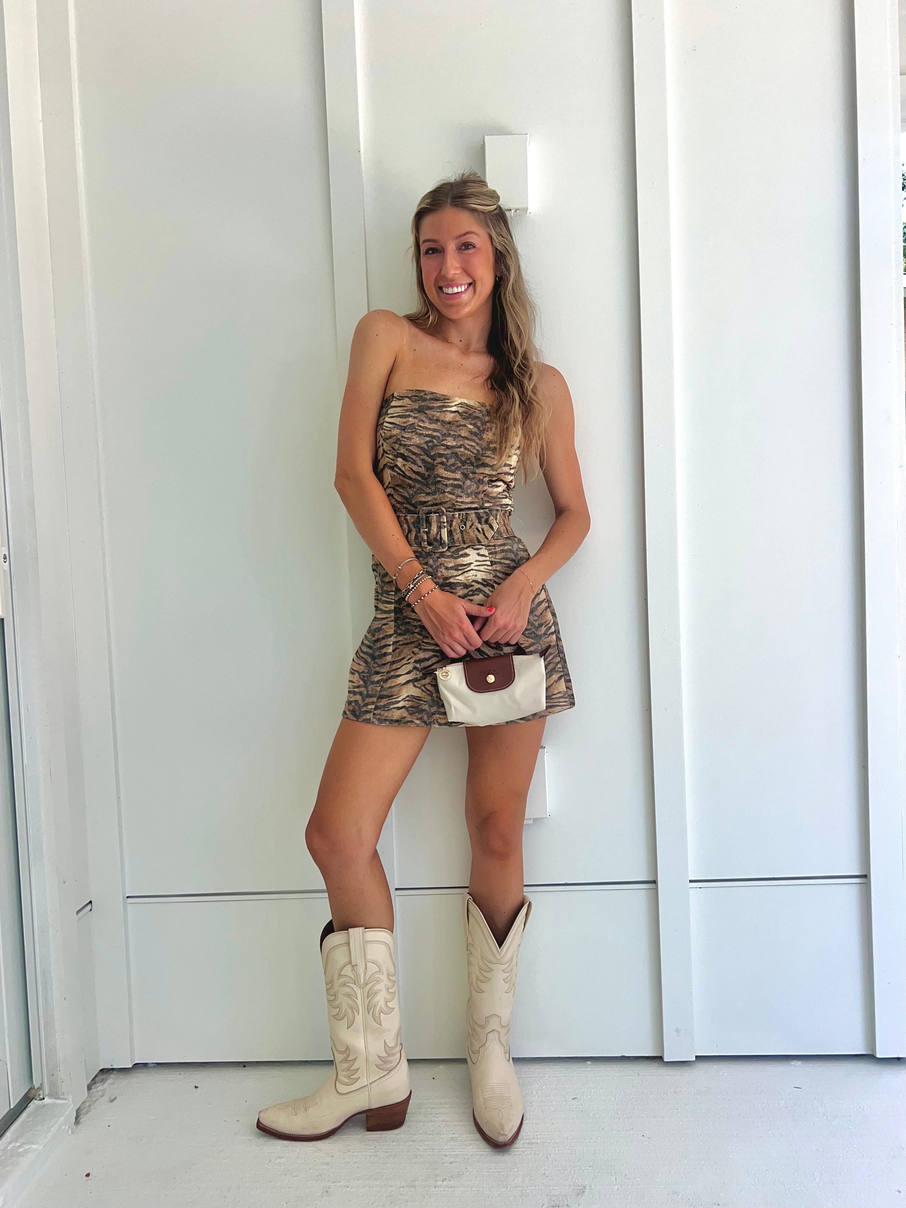 A woman wearing a strapless tiger printed dress and cream cowgirl boots with a cream purse standing against a white wall.