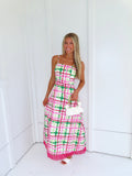 Woman wearing a pink and green plaid maxi dress holding a white purse against a minimal white wall.