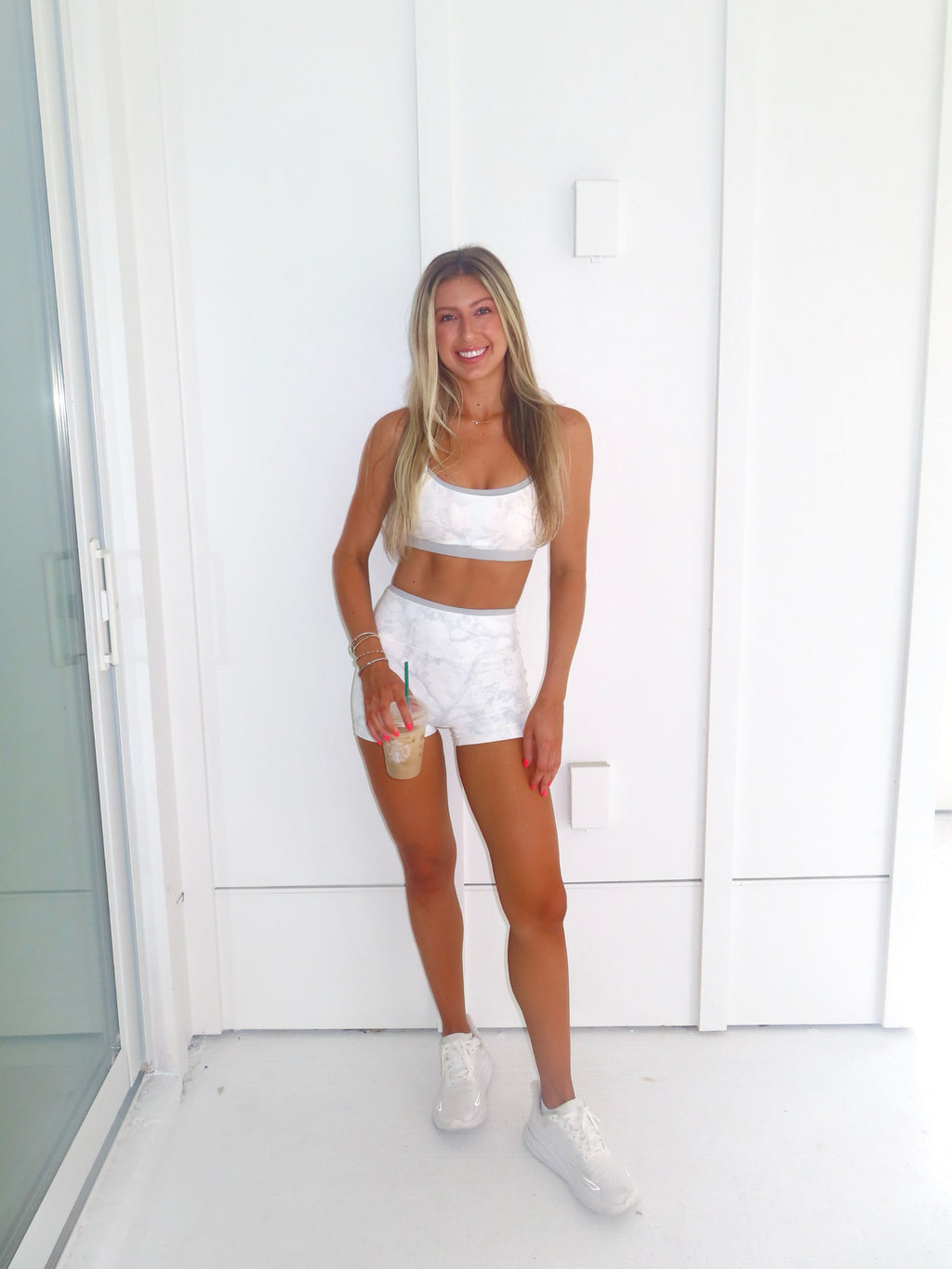 A woman stands against a white paneled wall wearing a white and grey camo sports bra and matching biker shorts, wearing white sneakers and holding a coffee.