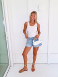 Woman wearing a white tank, denim shorts, and gold sandals holding a white purse against a white wall.