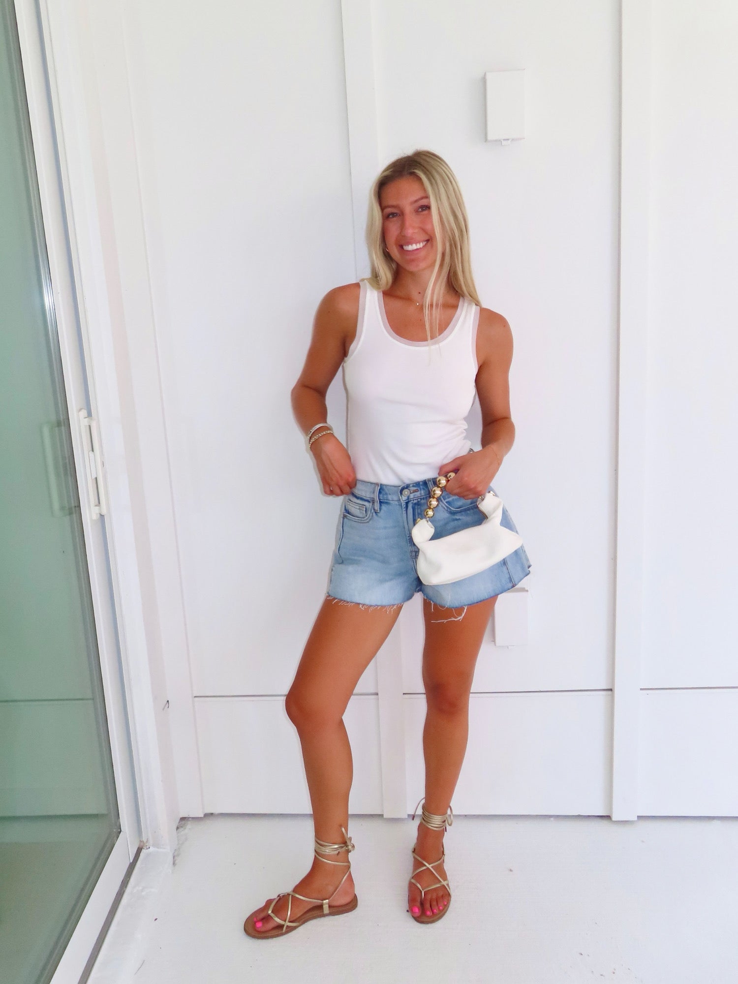 Woman wearing a white tank, denim shorts, and gold sandals holding a white purse against a white wall.