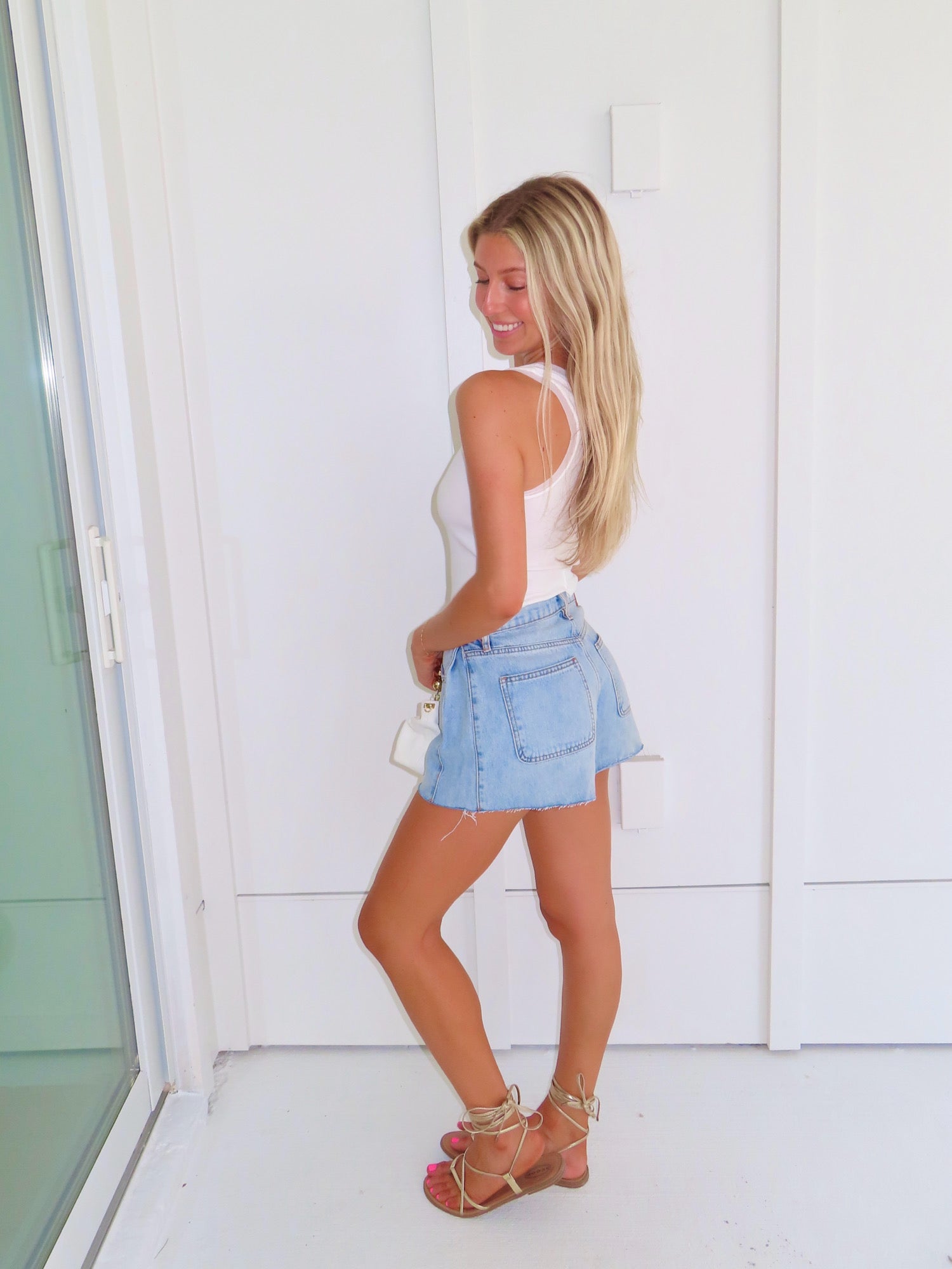 Woman wearing a white tank, denim shorts, and gold sandals holding a white purse against a white wall.