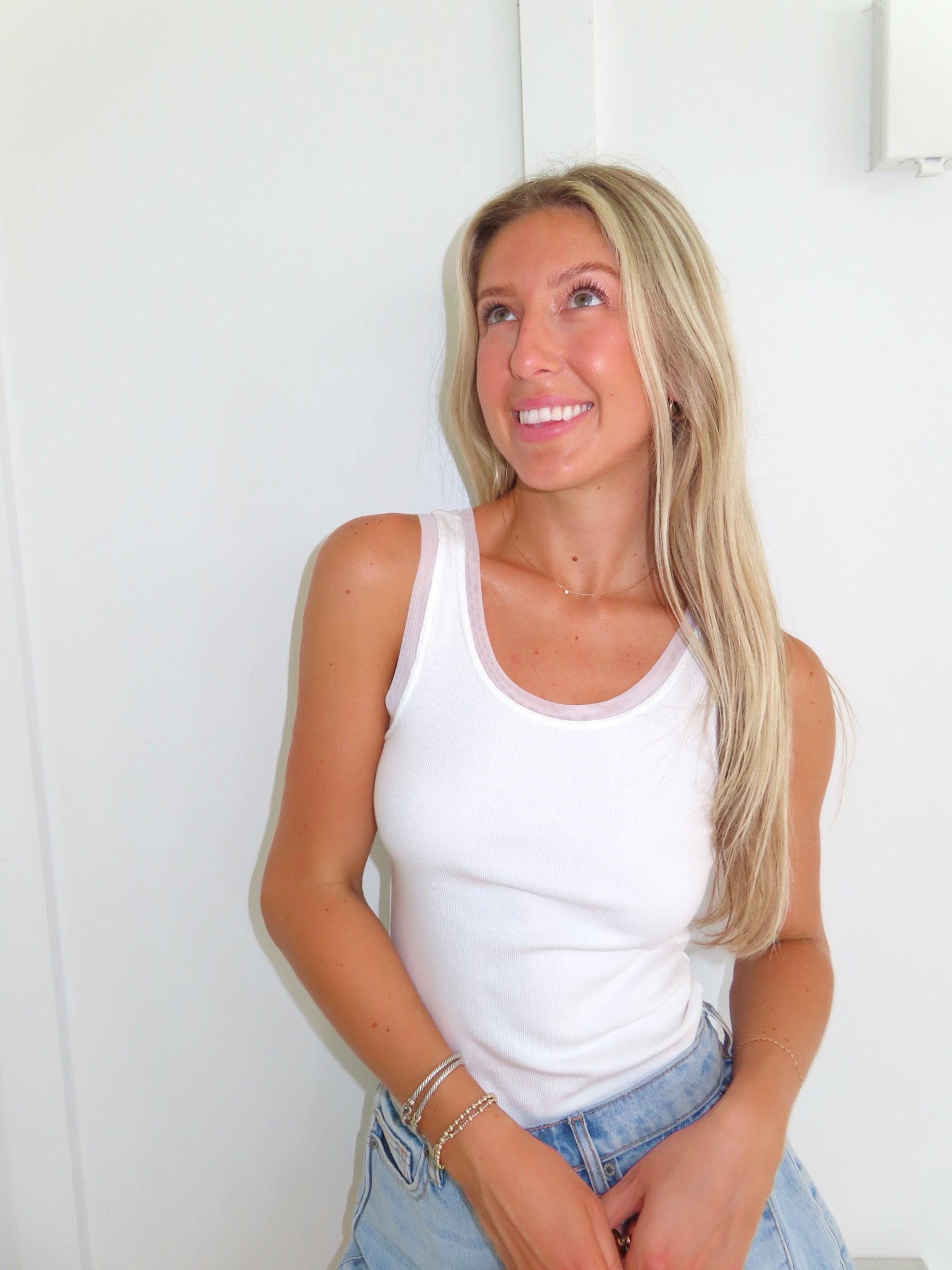 Woman wearing a white tank and denim shorts standing against a white wall.