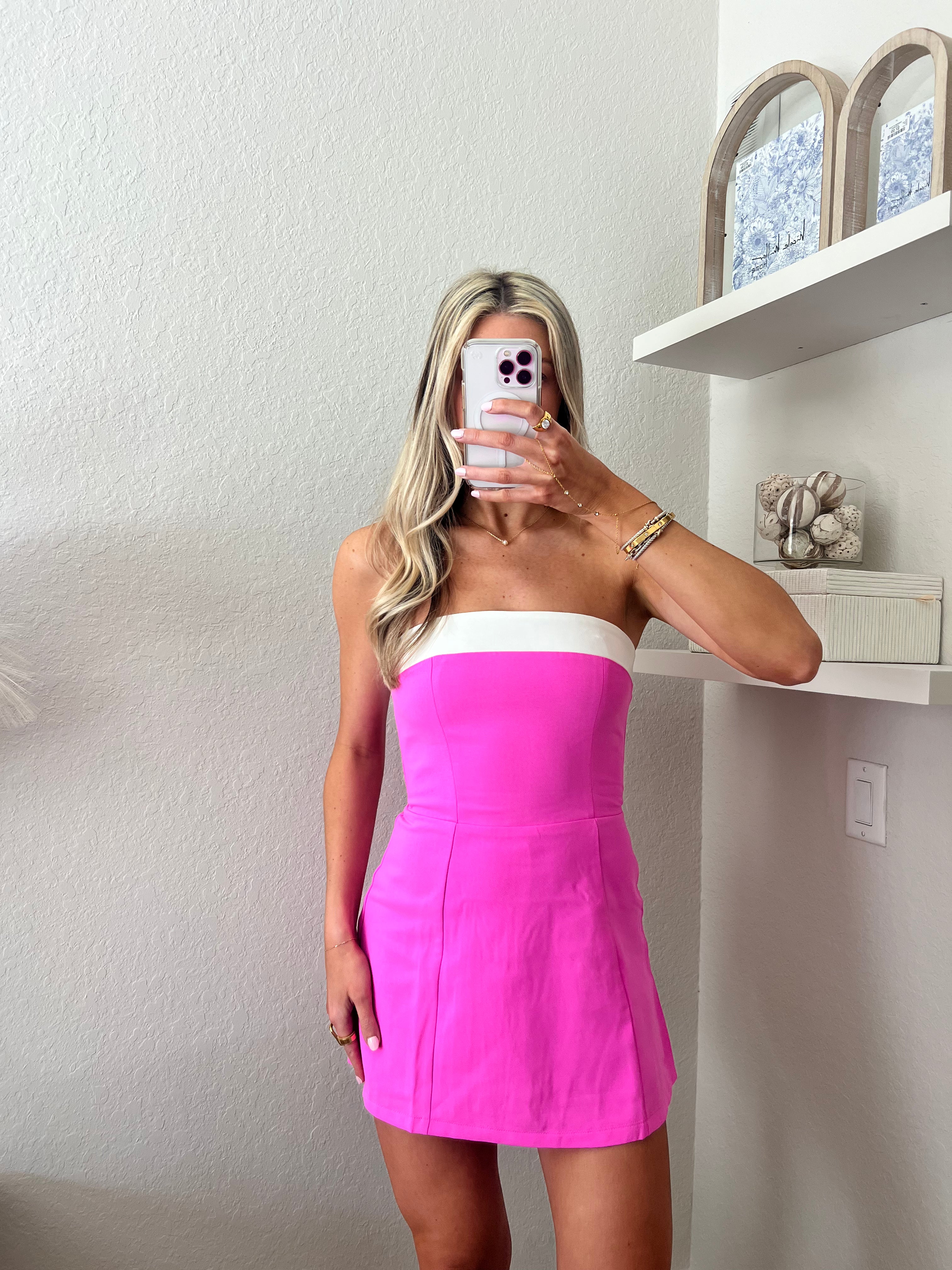 Woman taking a mirror selfie wearing a strapless pink mini dress with a white band at the top in a room.