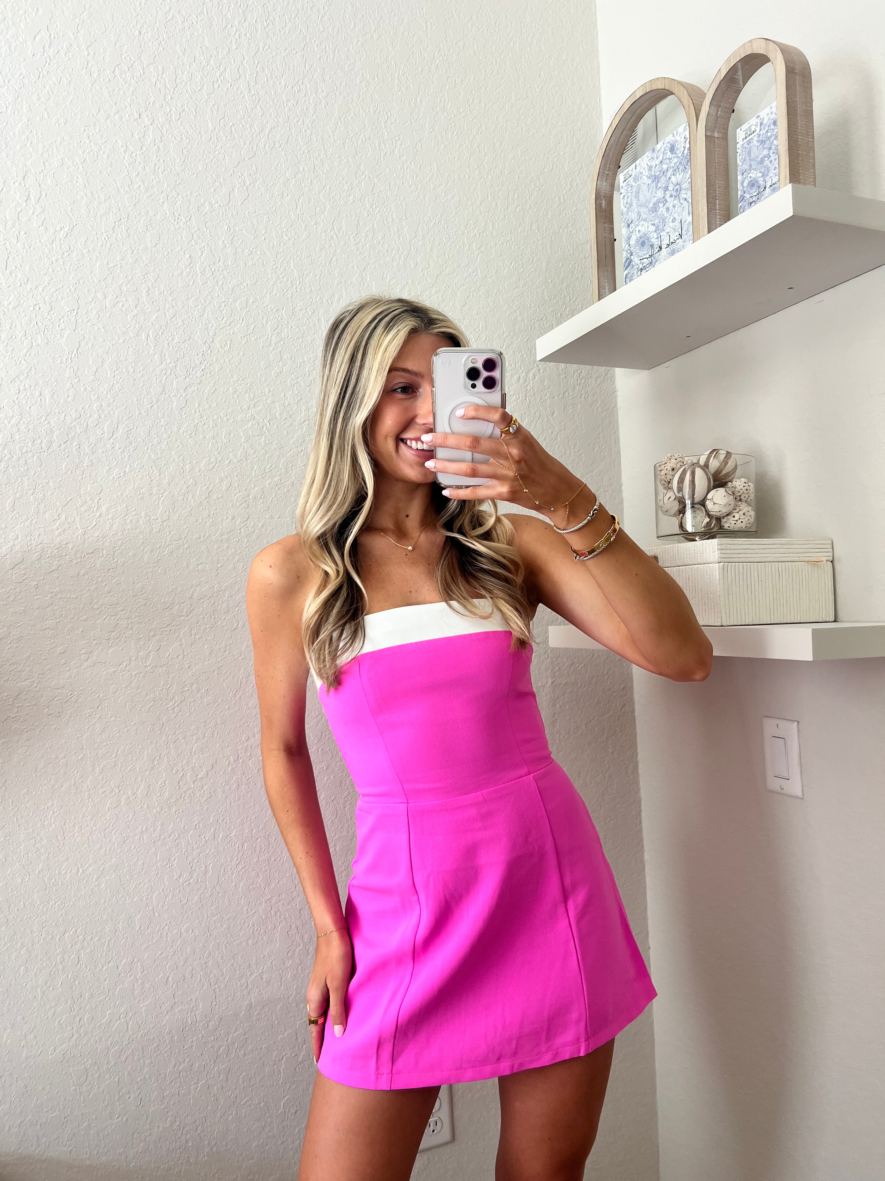 Woman taking a mirror selfie wearing a strapless pink mini dress with a white band at the top in a room.