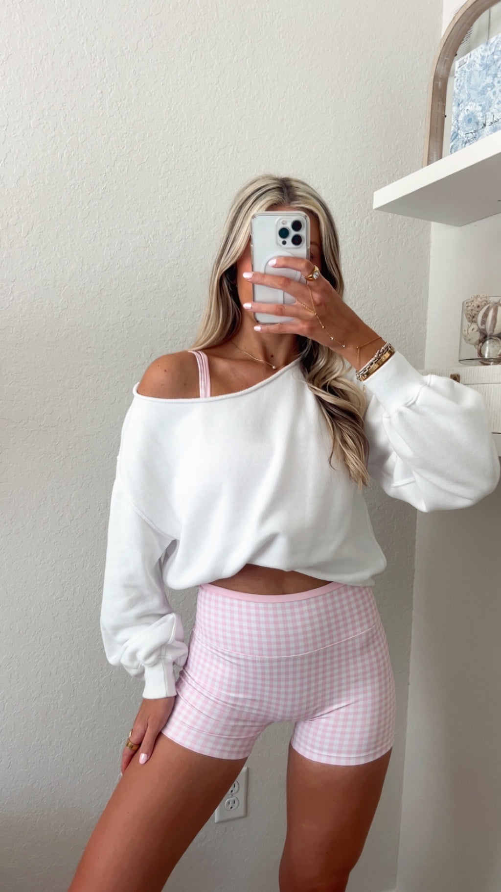 Woman taking a mirror selfie wearing a pink gingham, double layered sports bra and matching pink gingham biker shorts with a white off the shoulder top over.
