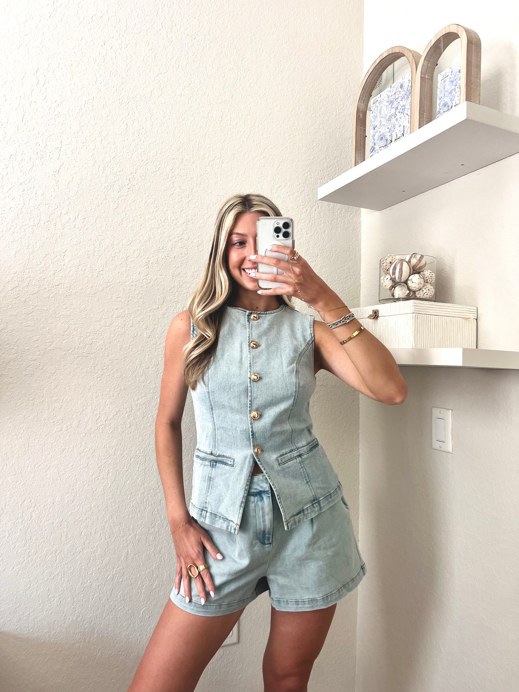 Woman taking a mirror selfie wearing a light blue denim top and shorts set with gold buttons.