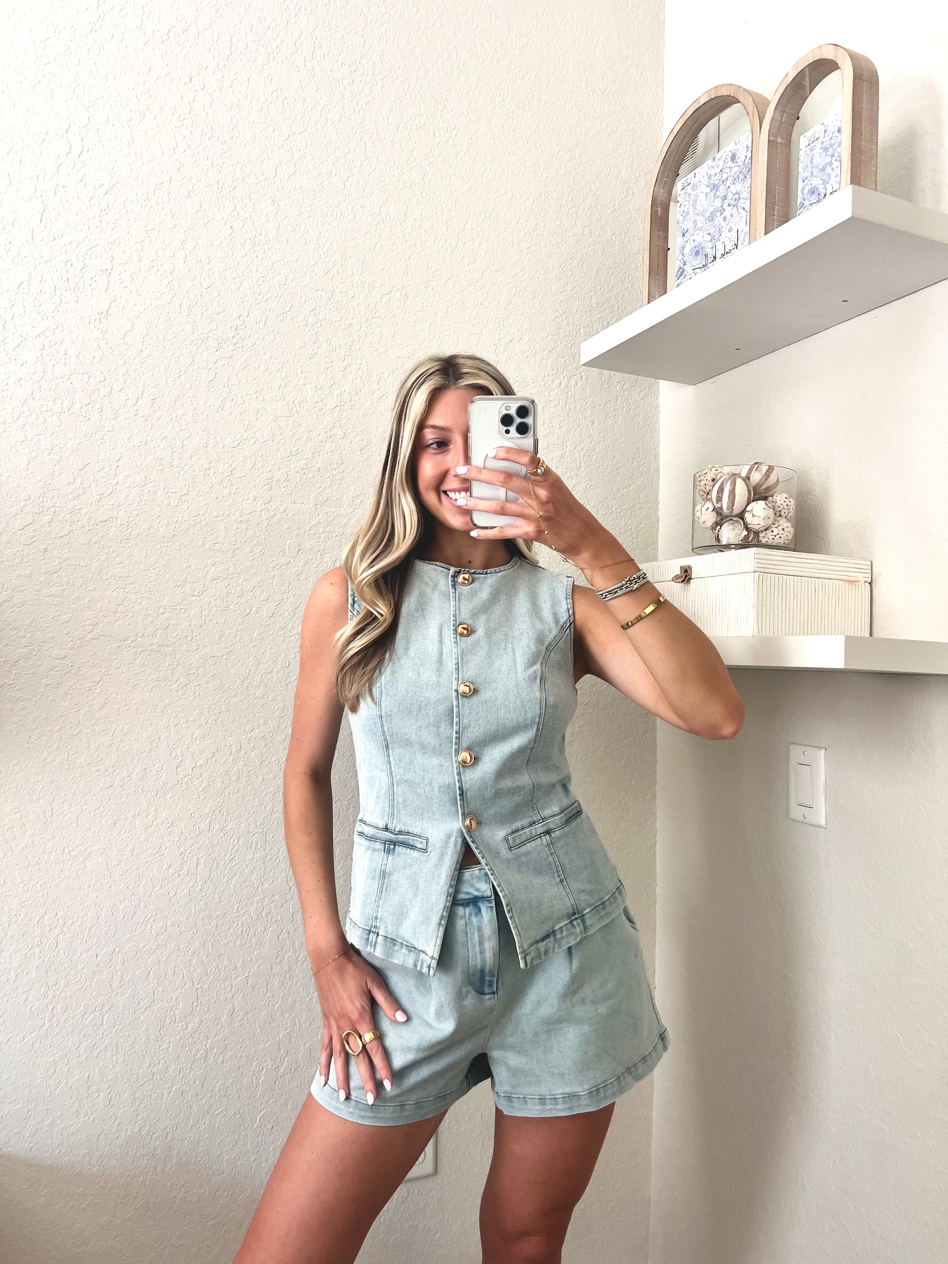 Woman taking a mirror selfie wearing a light blue denim top and shorts set with gold buttons.