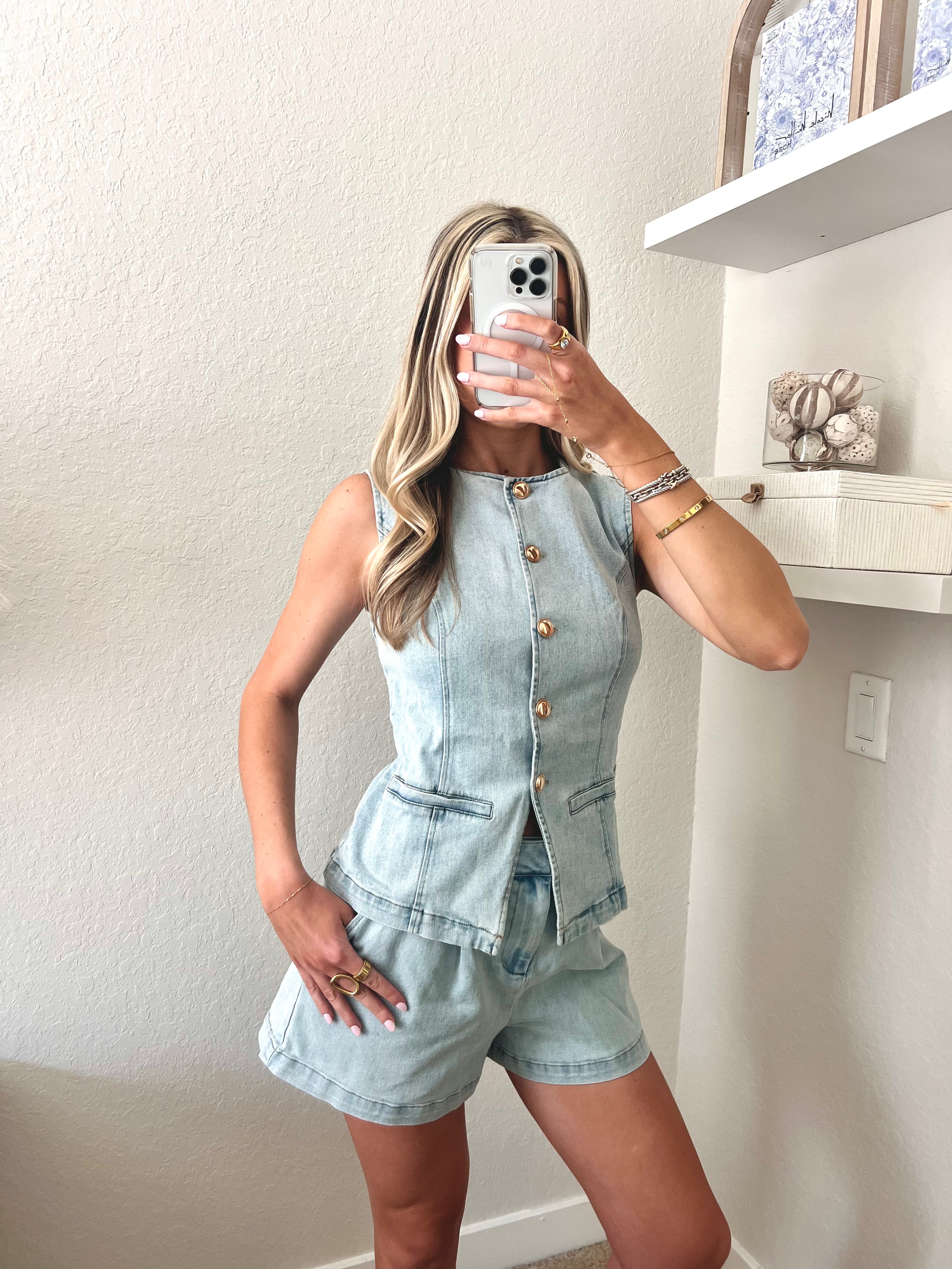 Woman taking a mirror selfie wearing a light blue denim top and shorts set with gold buttons.