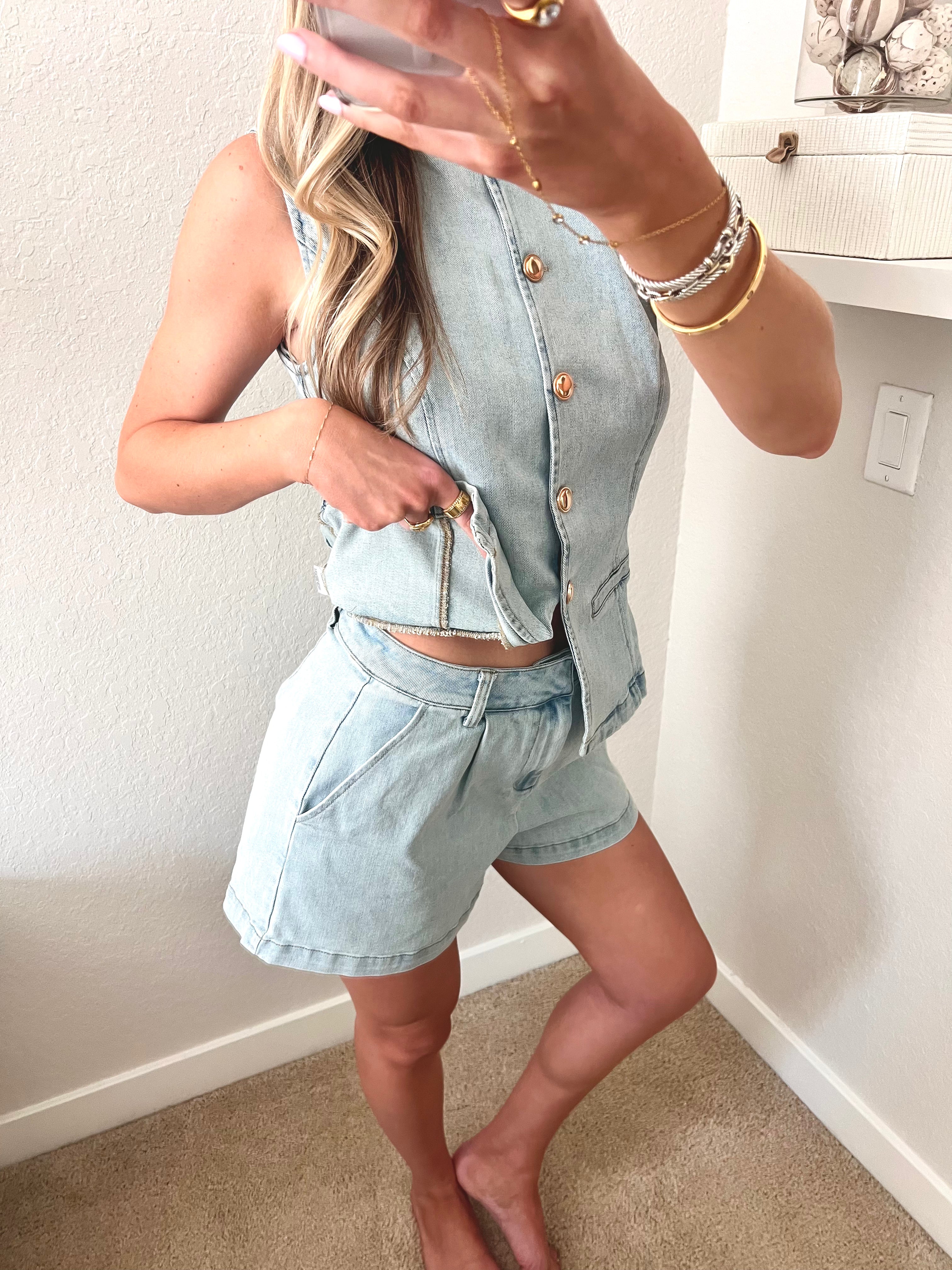 Woman taking a mirror selfie wearing a light blue denim top and shorts set with gold buttons.
