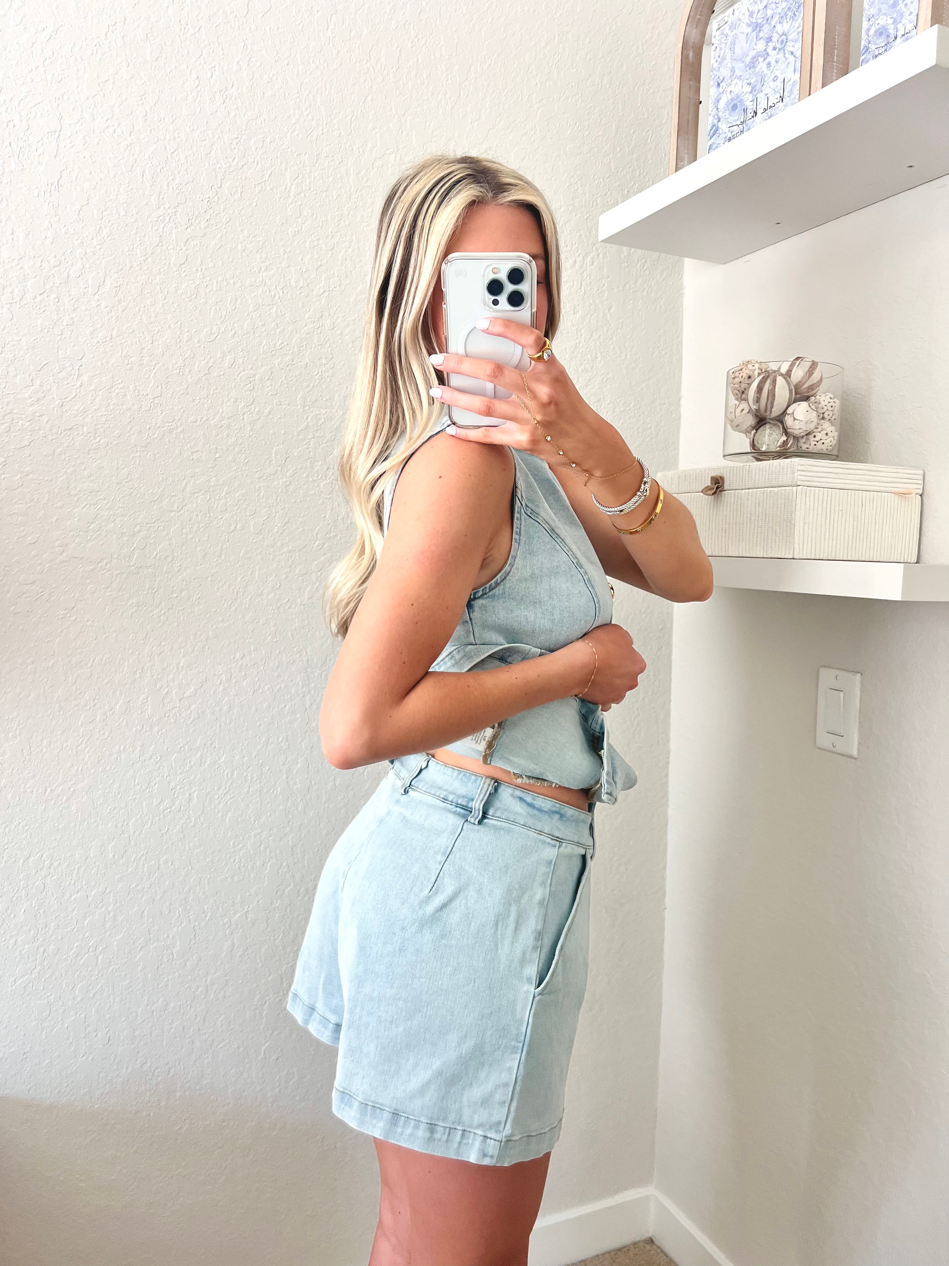 Woman taking a mirror selfie wearing a light blue denim top and shorts set with gold buttons.