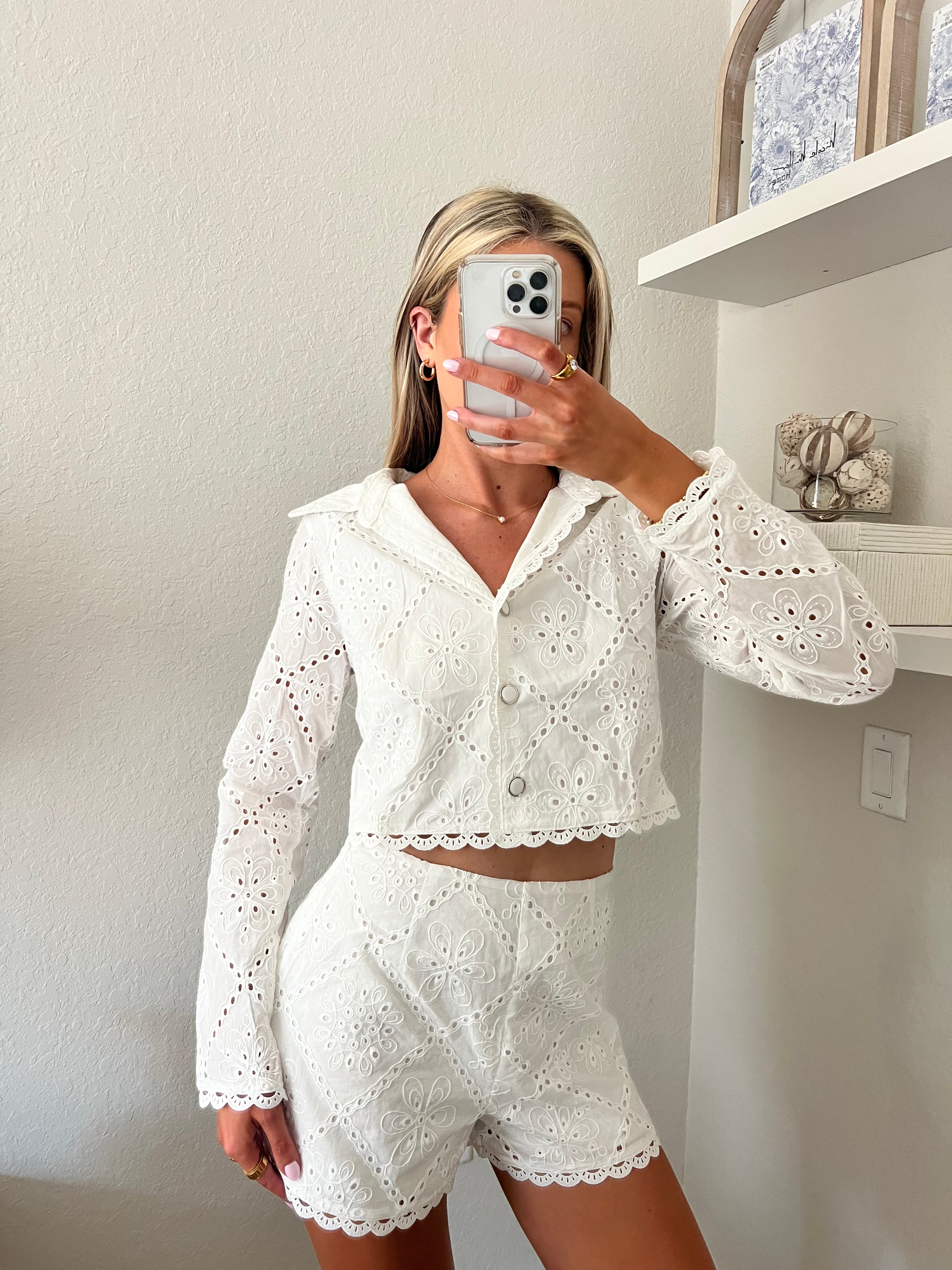 Woman taking a mirror selfie wearing a white lace outfit in a room with shelves. The outfit is a white lace long sleeve buttoned top with matching lace shorts.
