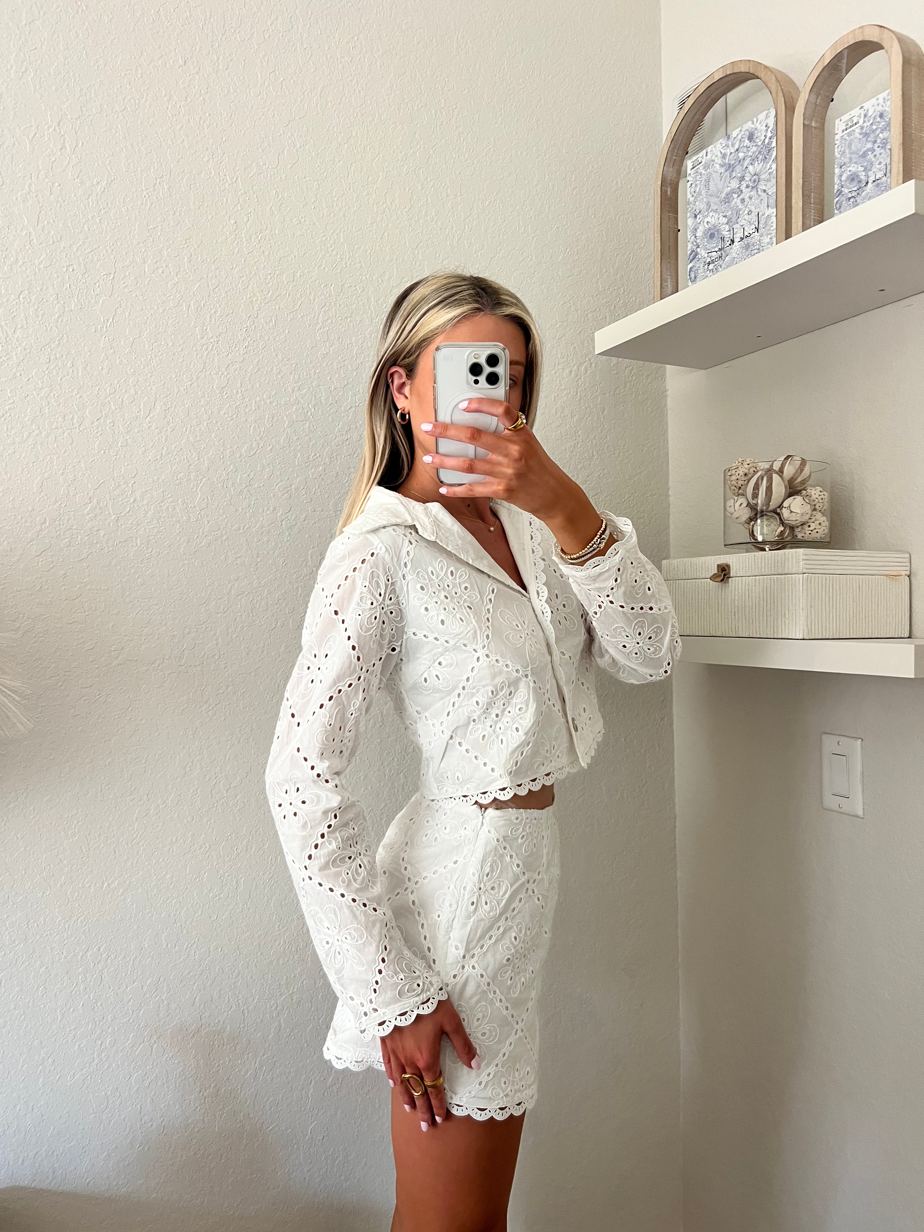 Woman taking a mirror selfie wearing a white lace outfit in a room with shelves. The outfit is a white lace long sleeve buttoned top with matching lace shorts.