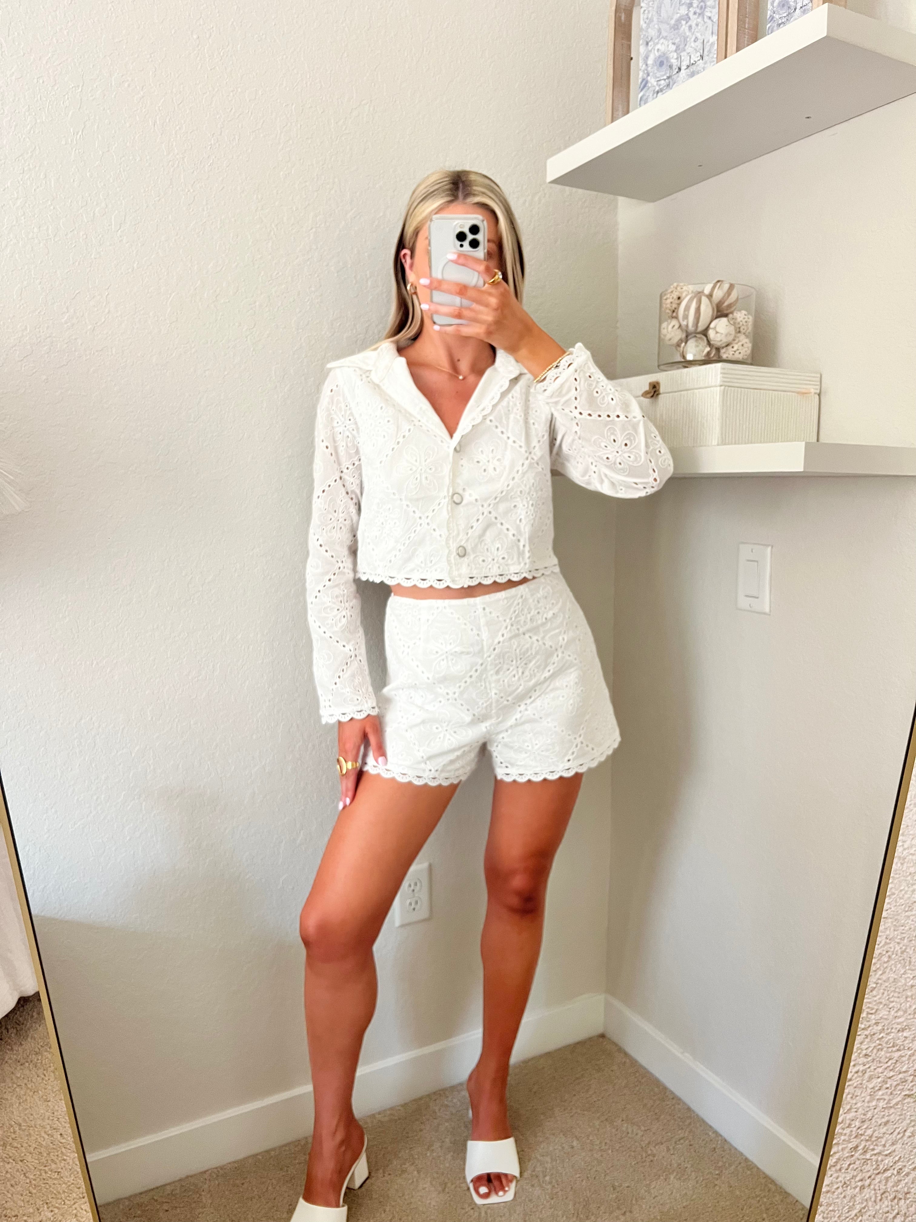 Woman taking a mirror selfie wearing a white lace outfit with white heels in a room with shelves. The outfit is a white lace long sleeve buttoned top with matching lace shorts.