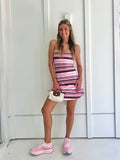A woman wearing a pink and brown striped athletic dress and pink sneakers with a cream purse standing against a white wall.