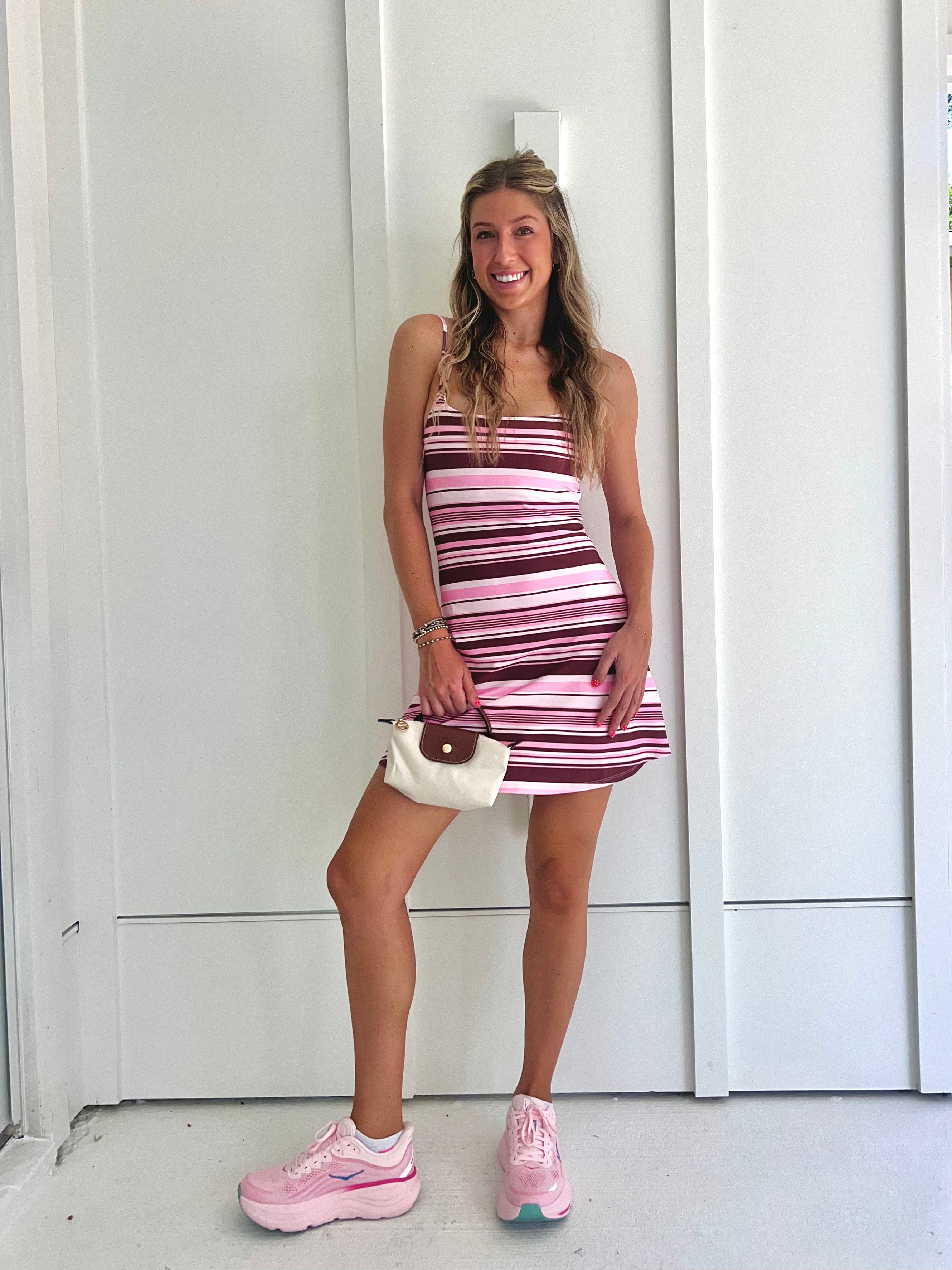 A woman wearing a pink and brown striped athletic dress and pink sneakers with a cream purse standing against a white wall.