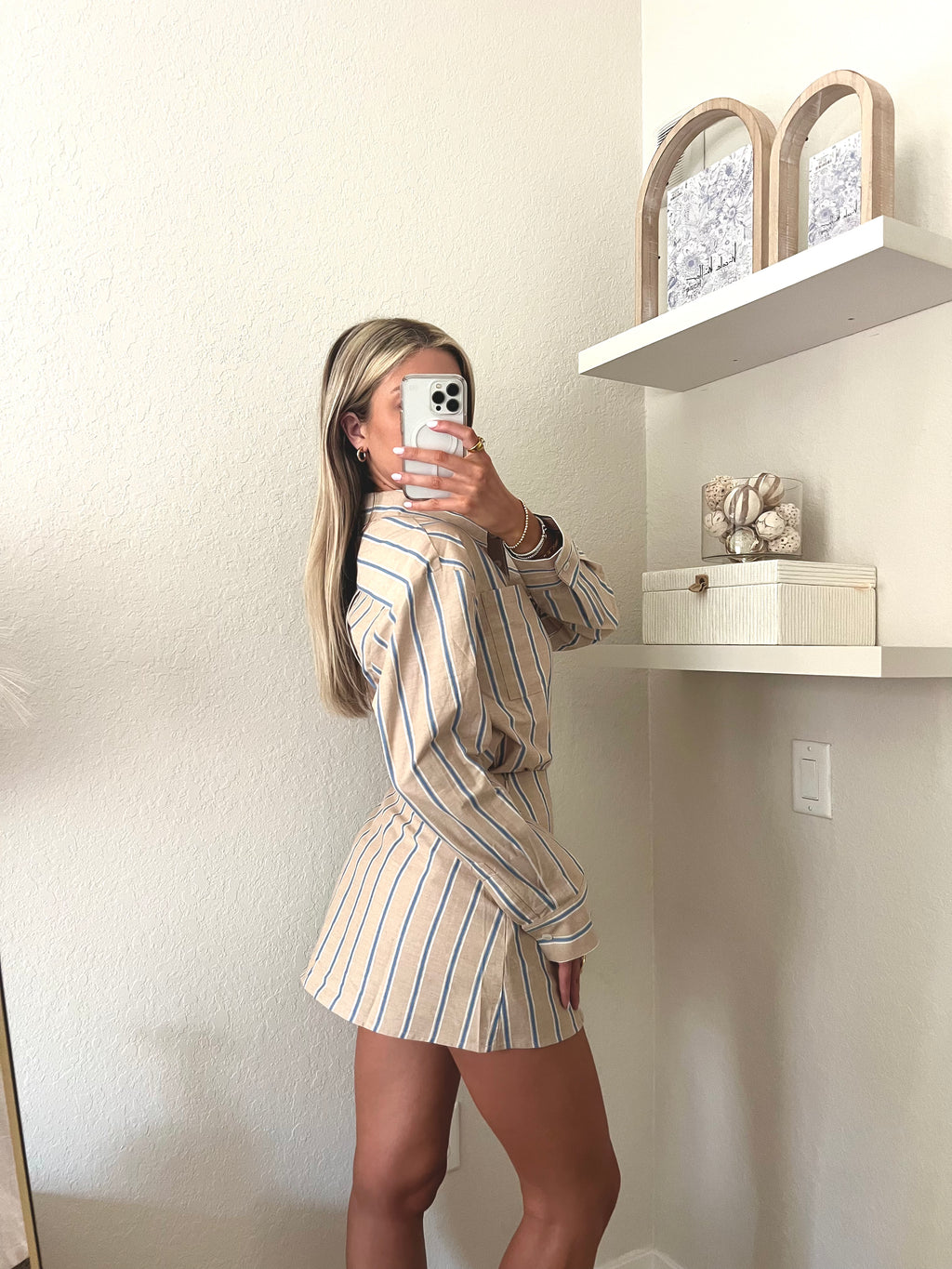 Woman taking a mirror selfie wearing a tan, blue, and white pin striped button down and skort set in a room with shelves and decor.