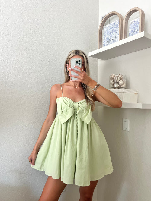 Woman taking a mirror selfie wearing a light green mini dress with a bow and button details.