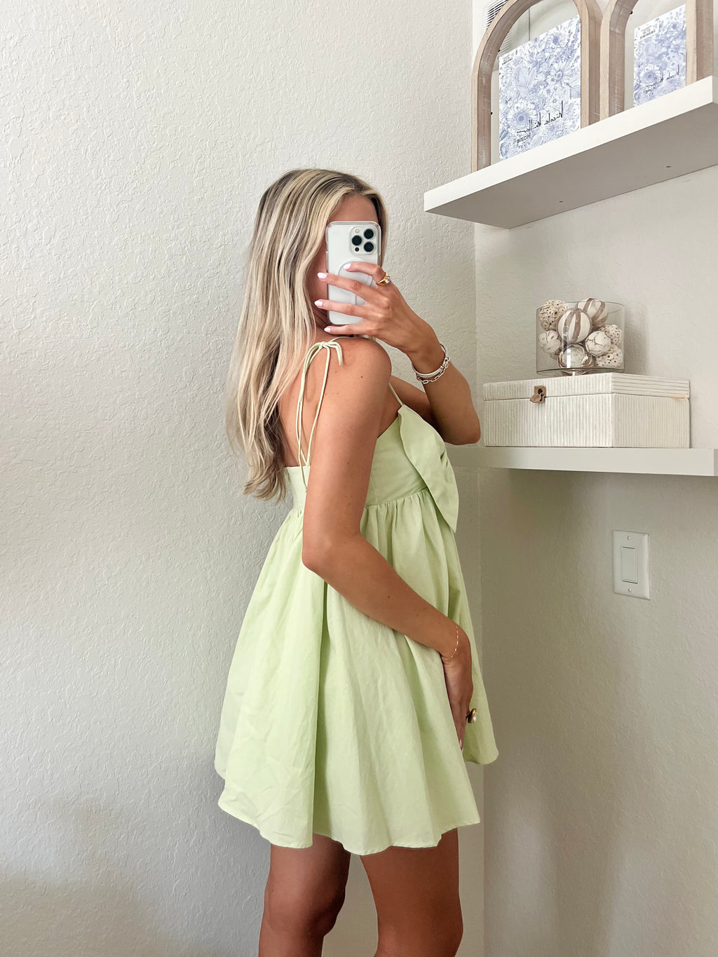 Woman taking a mirror selfie wearing a light green mini dress with a bow detail.