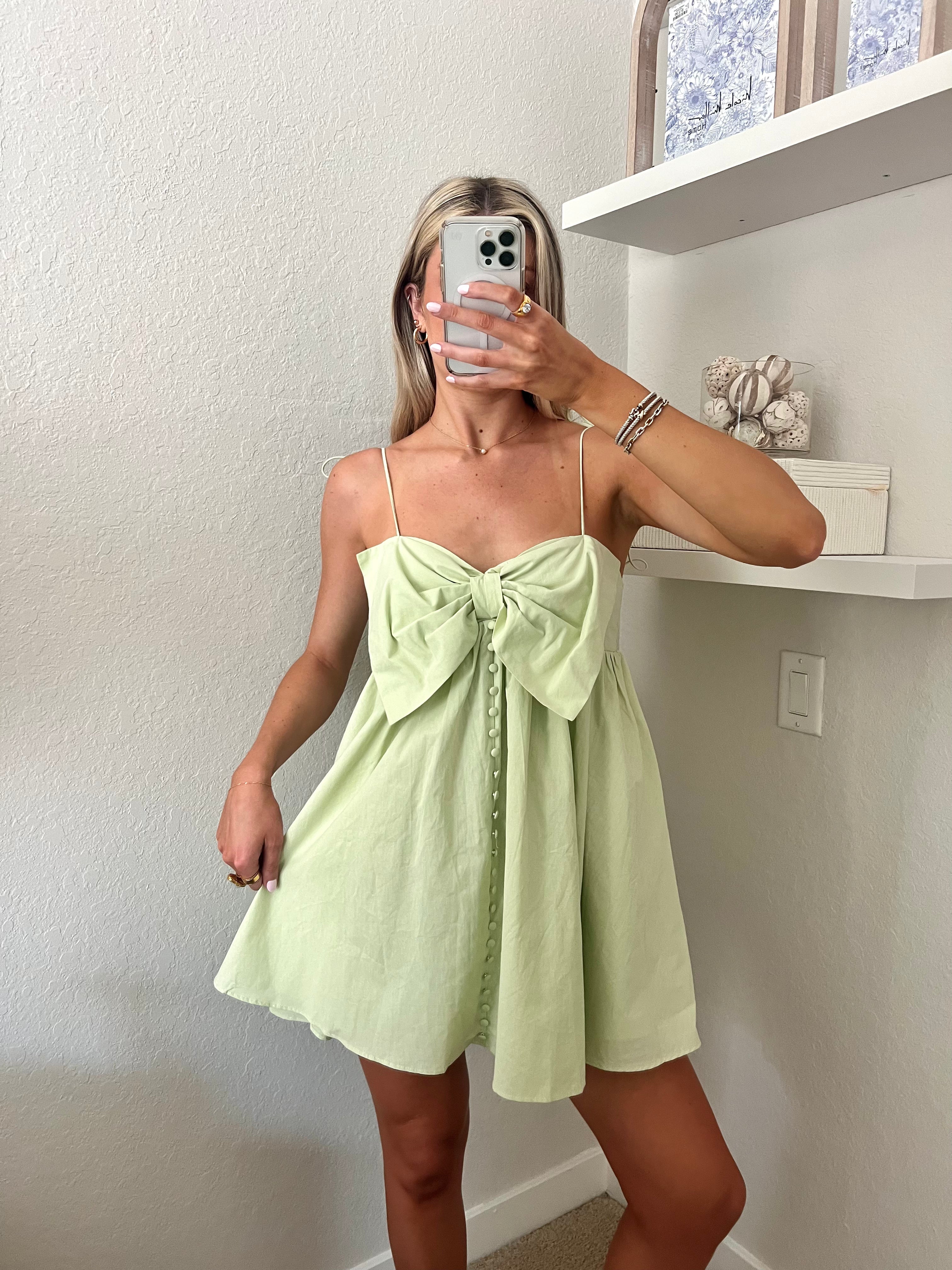 Woman taking a mirror selfie wearing a light green mini dress with a bow and button details.