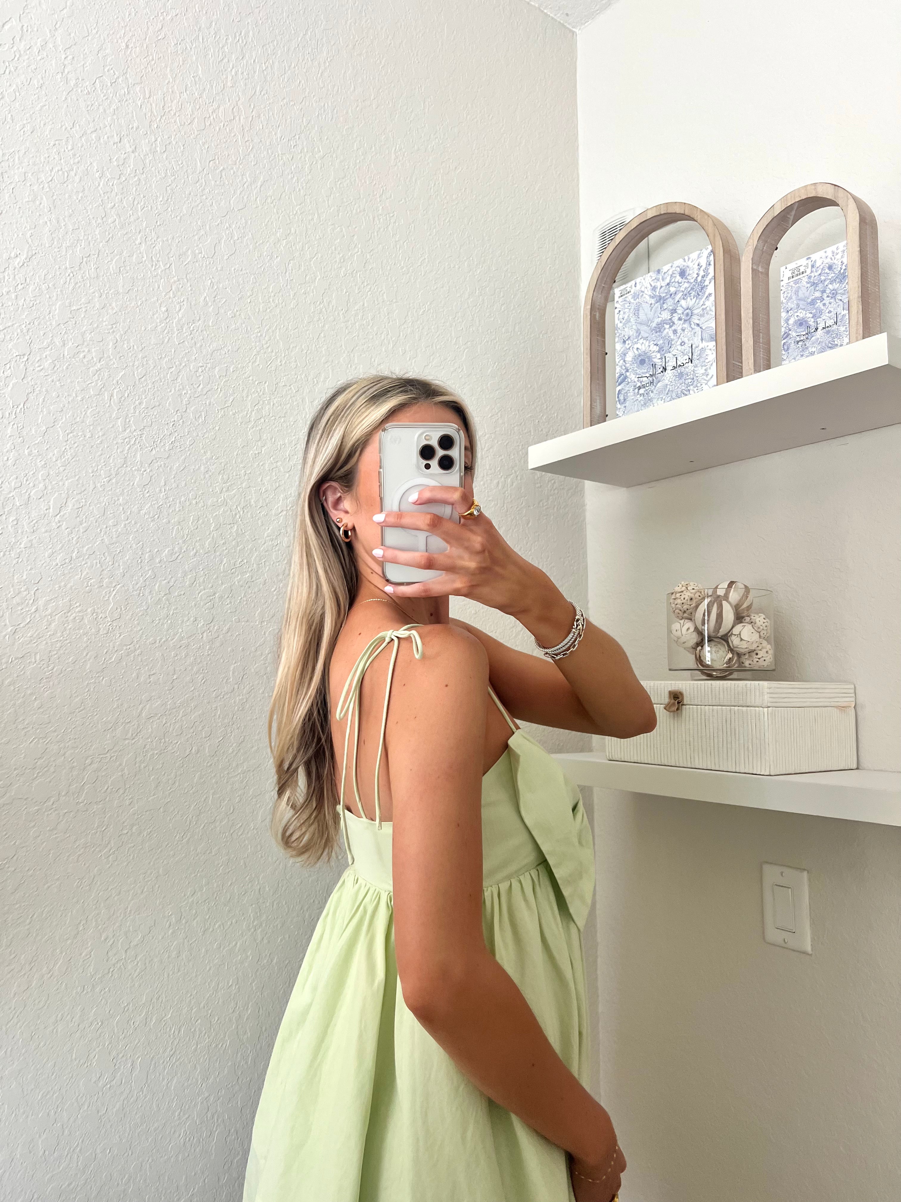 Woman taking a mirror selfie wearing a light green mini dress with a bow detail.