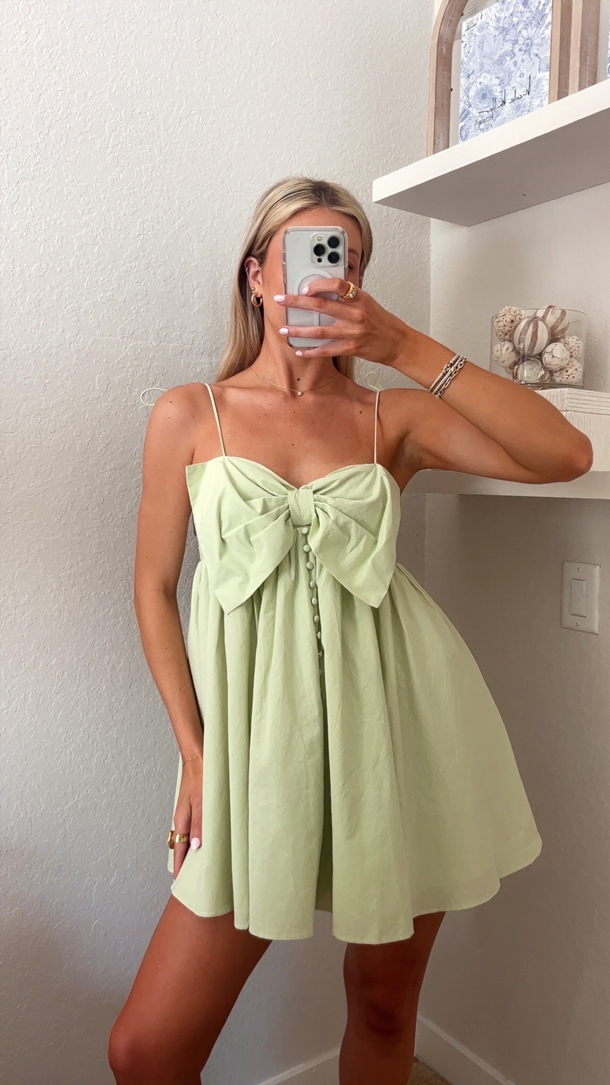 Woman taking a mirror selfie wearing a light green mini dress with a bow and button details.