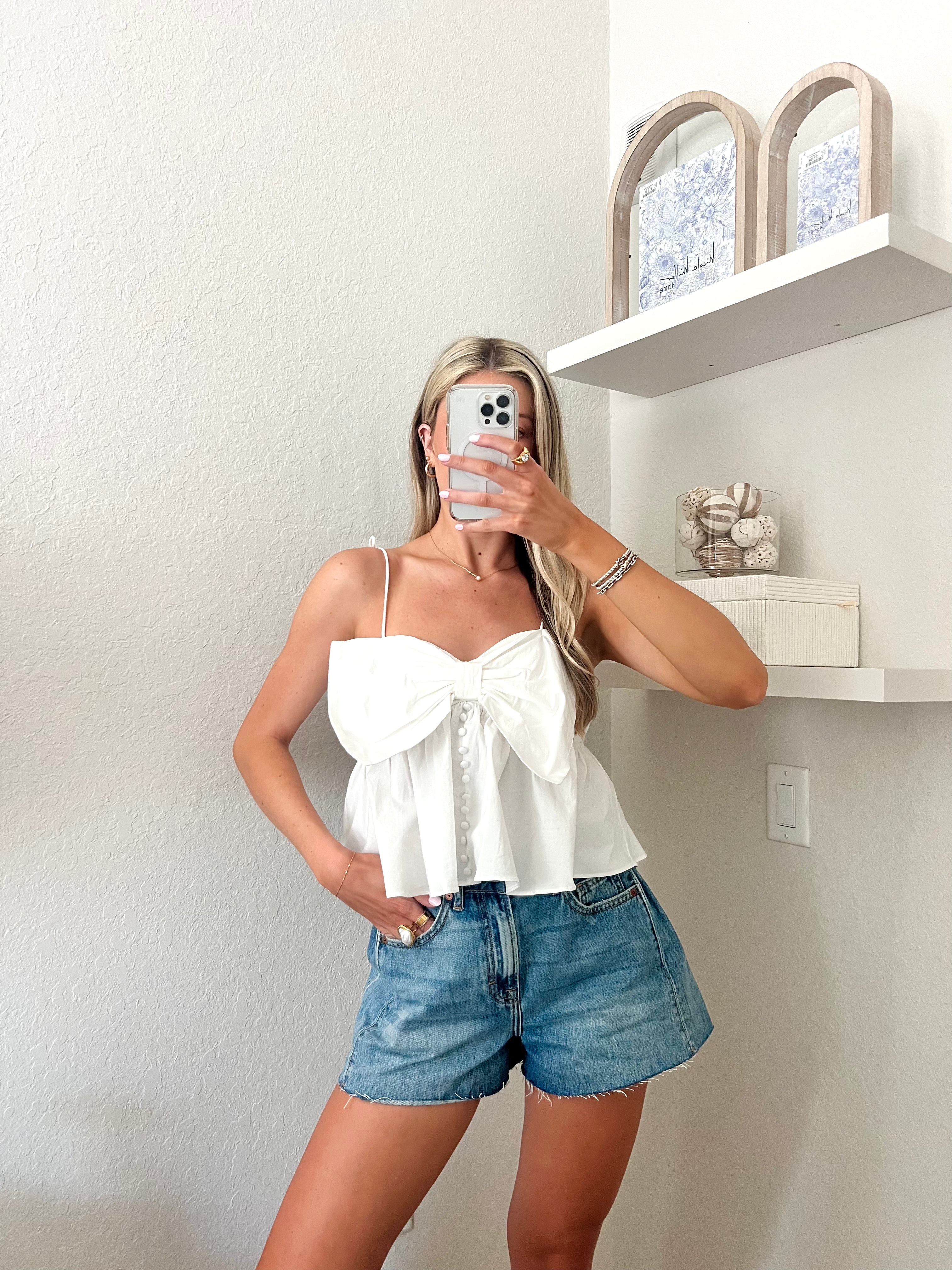 Woman taking a mirror selfie wearing a white top and blue denim shorts in a minimalistic room.
