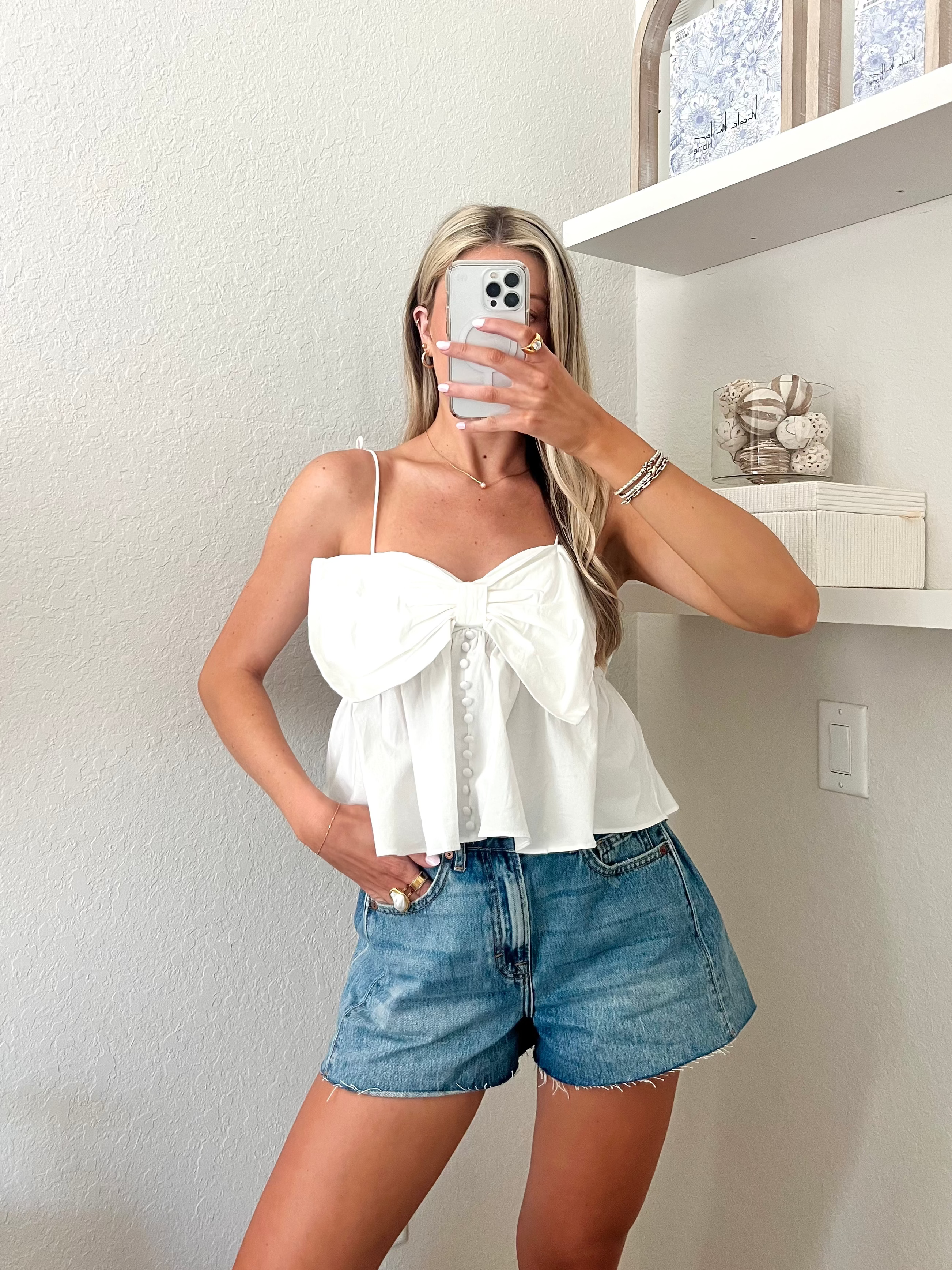 Woman taking a mirror picture wearing a white top with a bow detail and blue jean shorts.