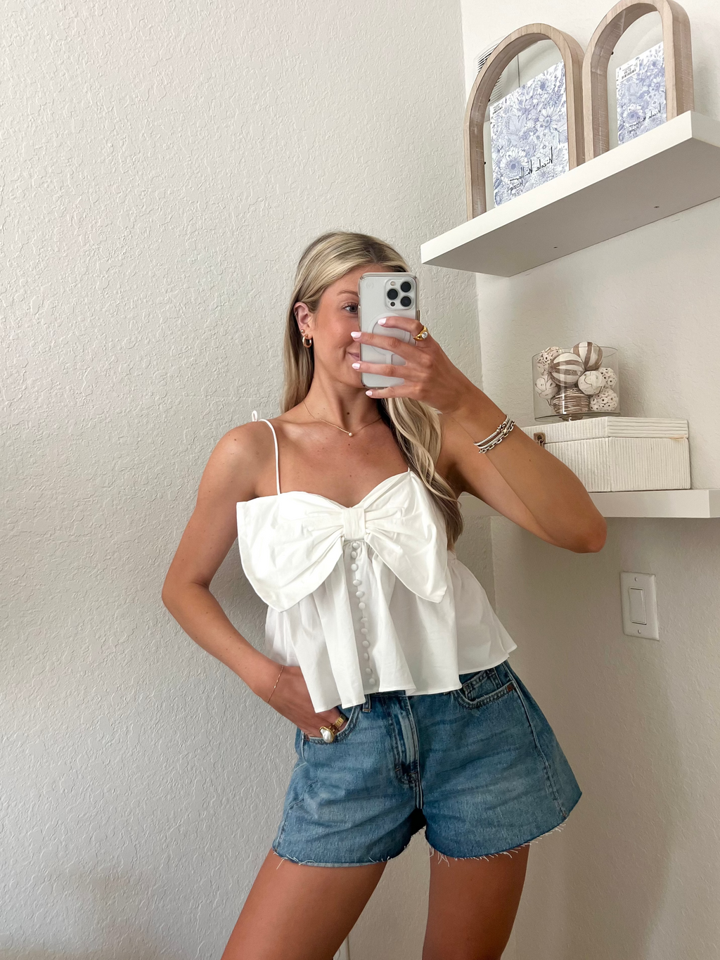 Woman taking a mirror picture wearing a white top with a bow detail and blue jean shorts.