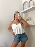 Woman taking a mirror picture wearing a white top with a bow detail and blue jean shorts.