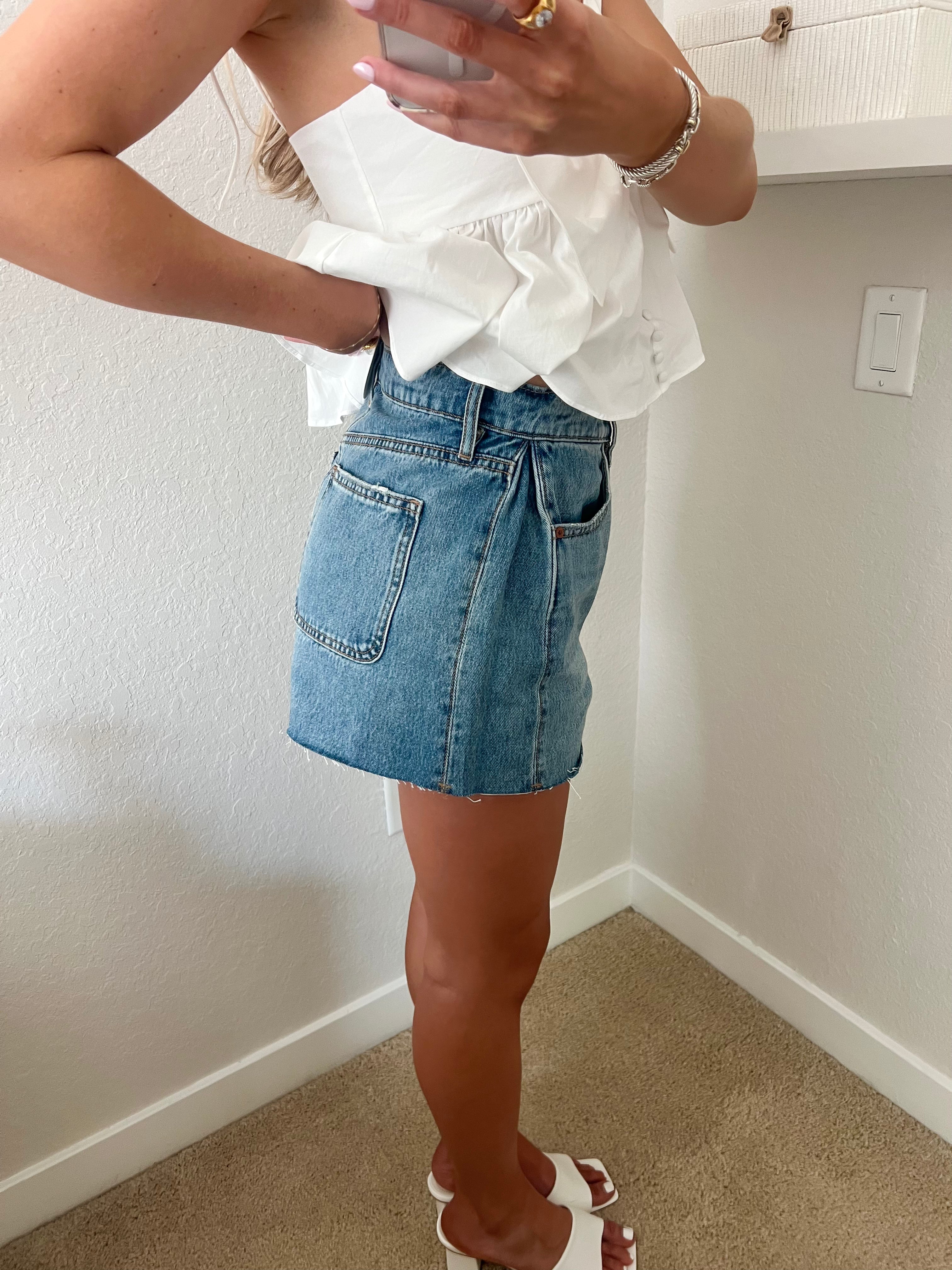 Woman taking a mirror selfie wearing a white top and blue denim shorts with white heels standing against a plain wall.