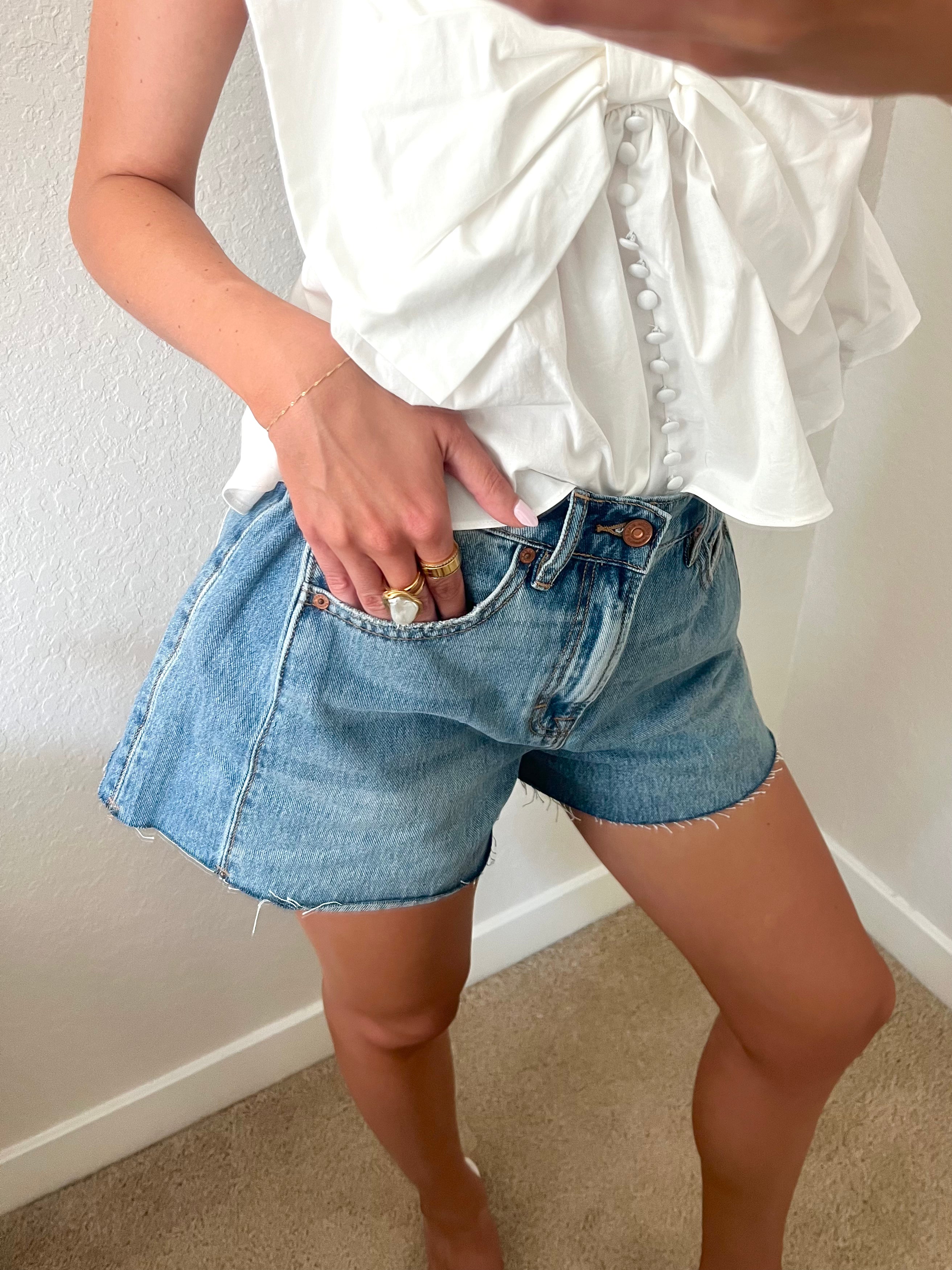 Woman taking a mirror selfie wearing a white top and blue denim shorts standing against a plain wall.