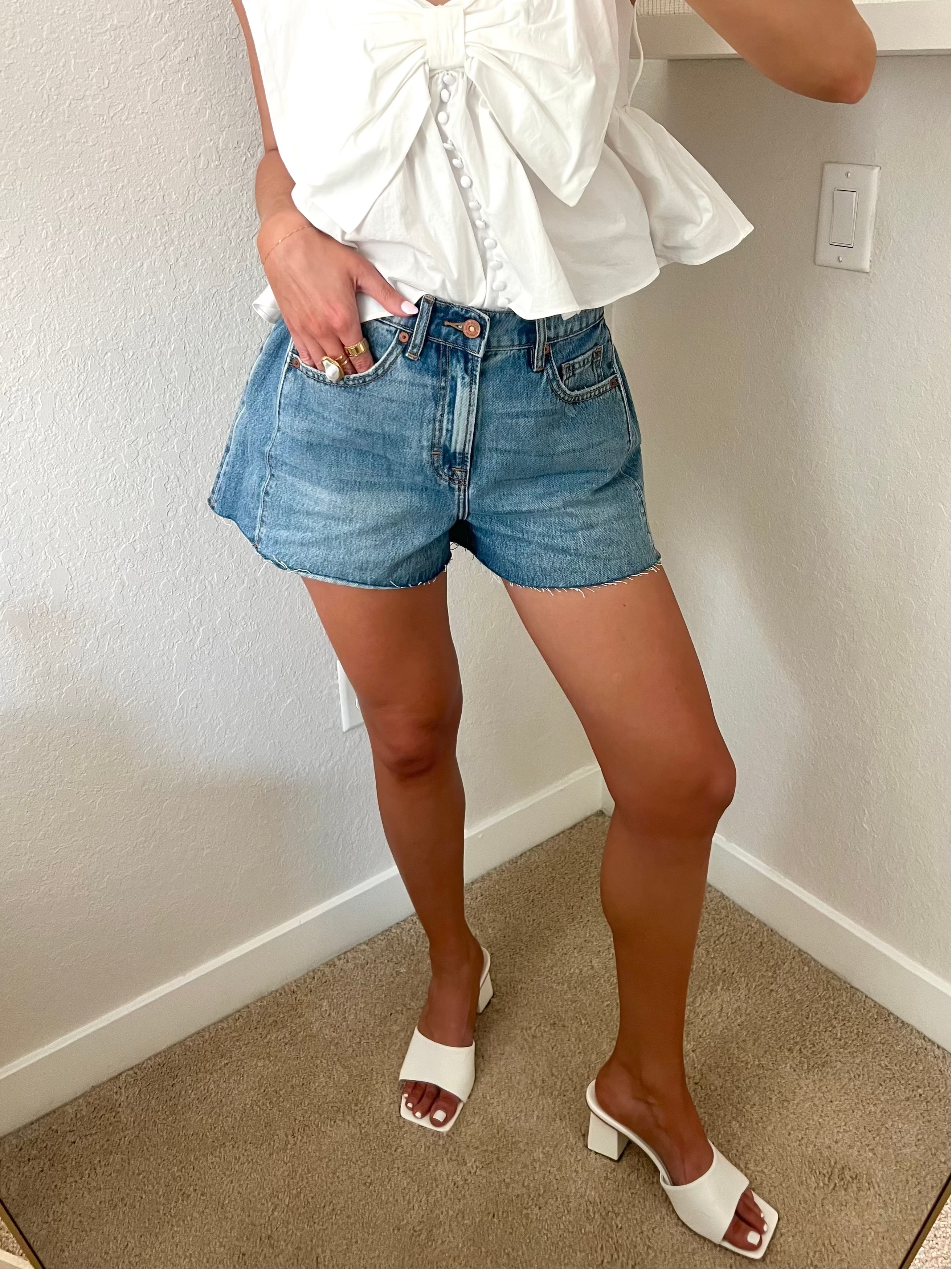 Woman taking a mirror selfie wearing a white top and blue denim shorts standing against a plain wall.
