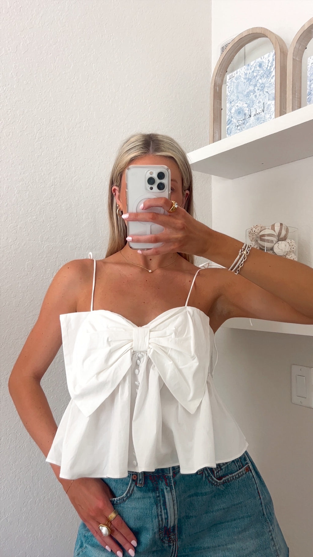 Woman taking a mirror picture wearing a white top with a bow detail and blue jean shorts.
