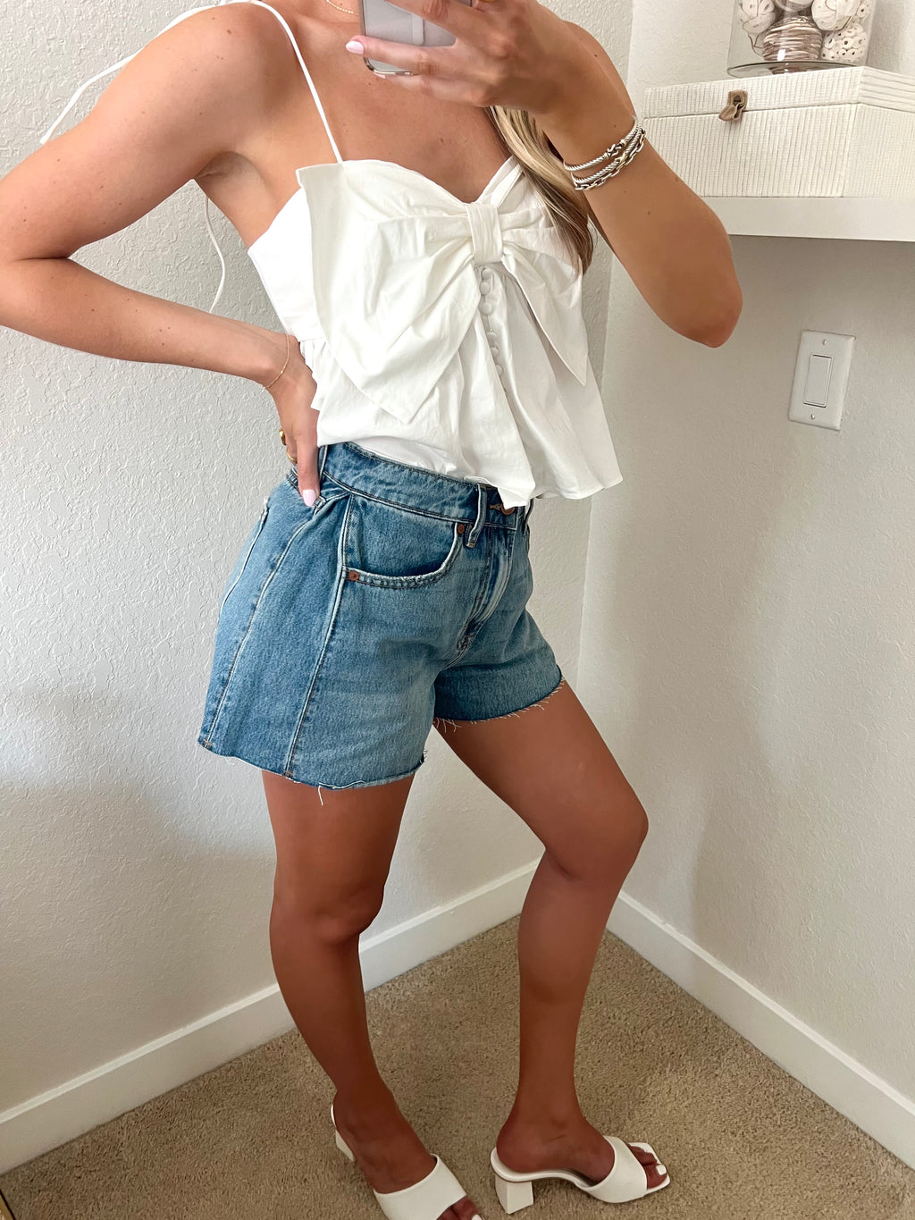 Woman taking a mirror selfie wearing a white top and blue denim shorts with white heels standing against a plain wall.