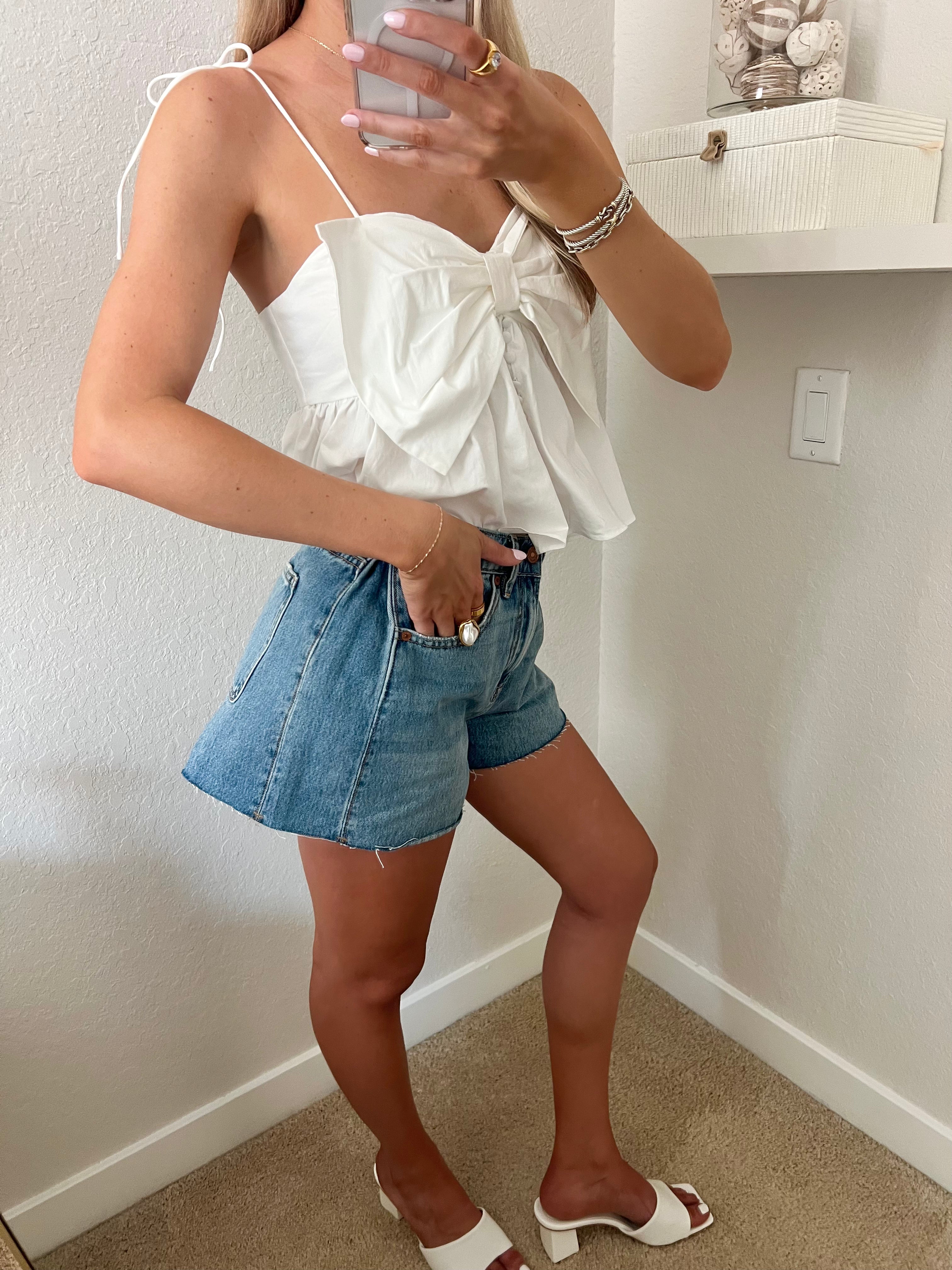 Woman taking a mirror selfie wearing a white top and blue denim shorts with white heels standing against a plain wall.