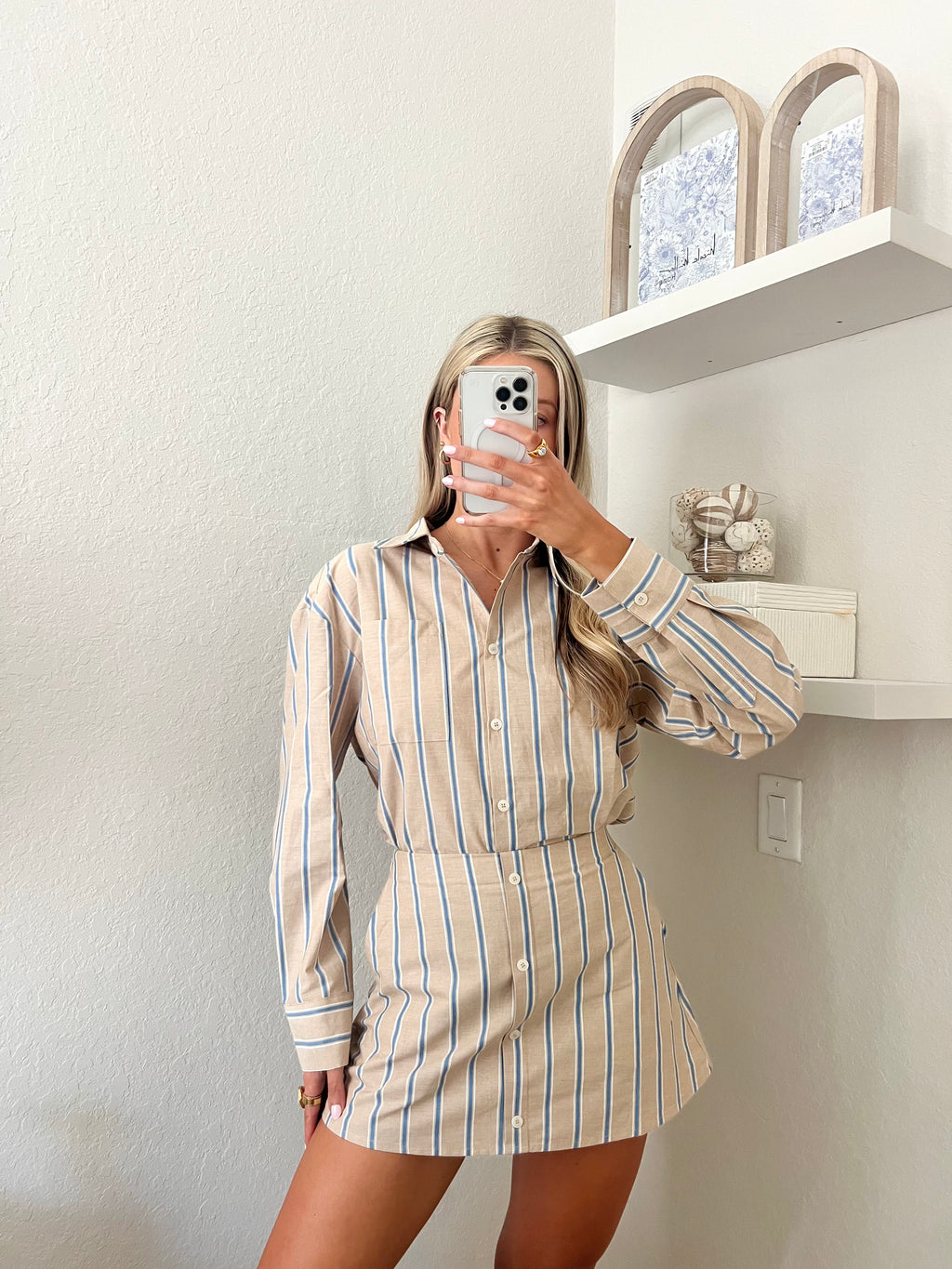 Woman taking a mirror selfie wearing a tan, blue, and white pin striped button down and skort set in a room with shelves and decor.