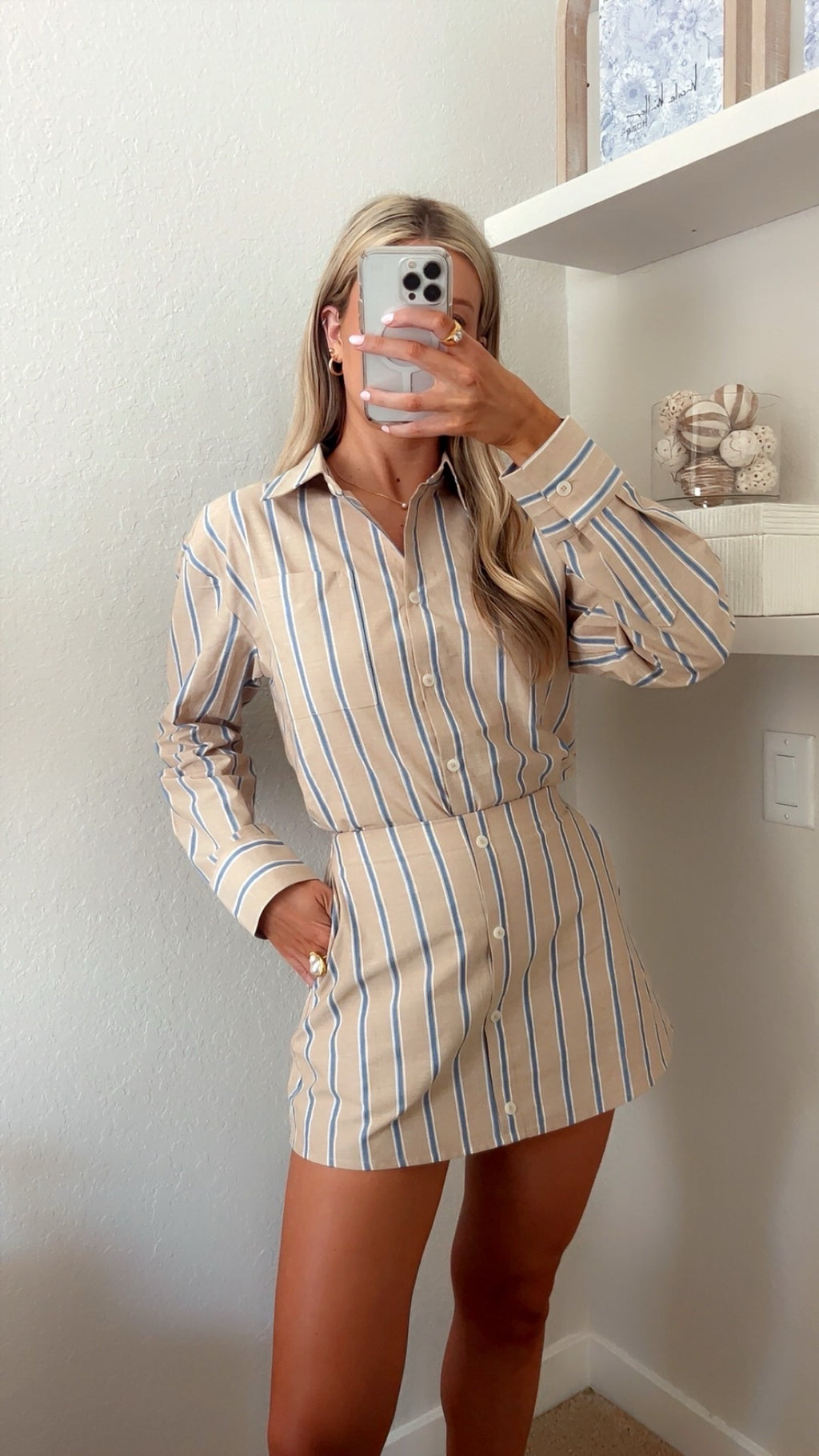 Woman taking a mirror selfie wearing a tan, blue, and white pin striped button down and skort set in a room with shelves and decor.