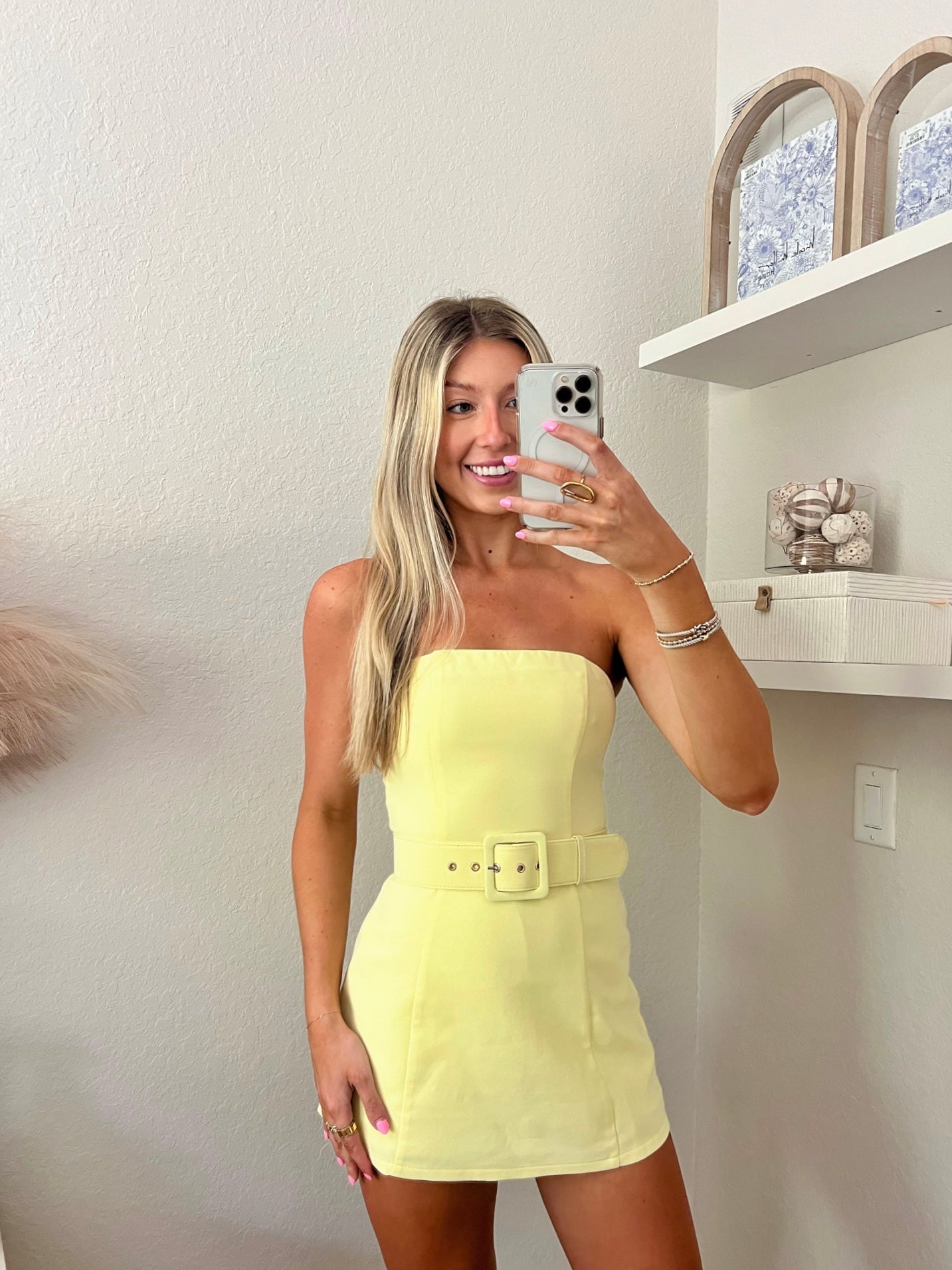 Woman taking a mirror selfie wearing a yellow strapless belted dress in a minimalist room.