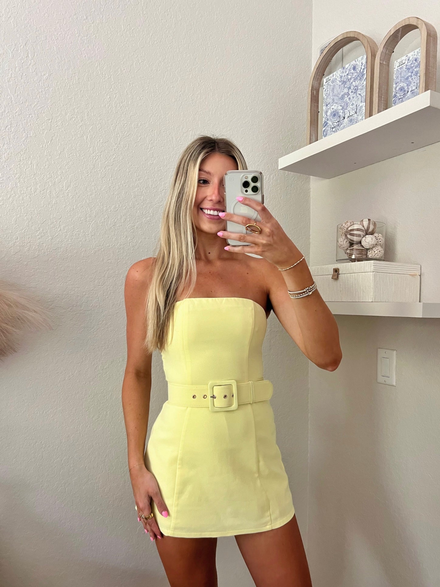 Woman taking a mirror selfie wearing a yellow strapless belted dress in a minimalist room.