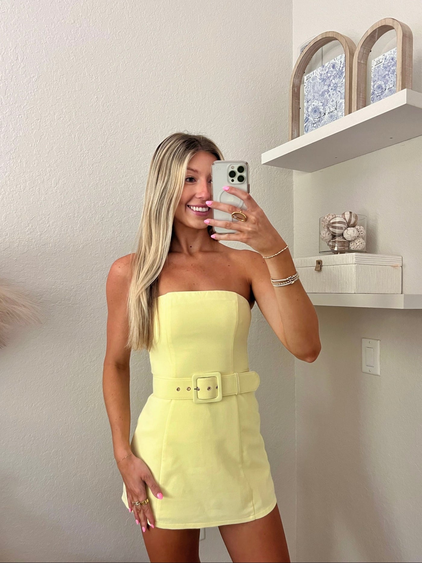 Woman taking a mirror selfie wearing a yellow strapless belted dress in a minimalist room.