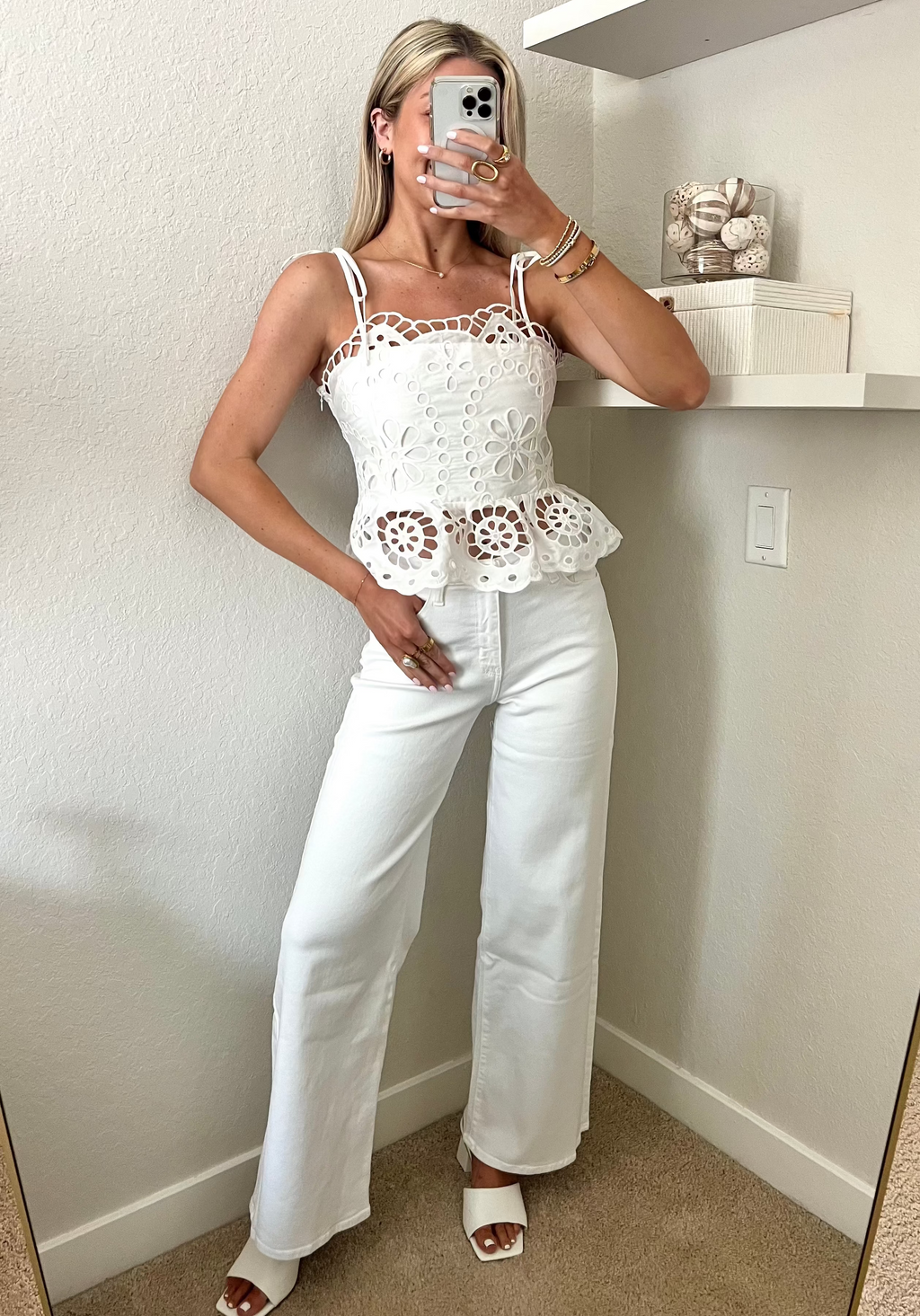 Woman taking a mirror selfie wearing a white lace top and white jeans with heels in a room.