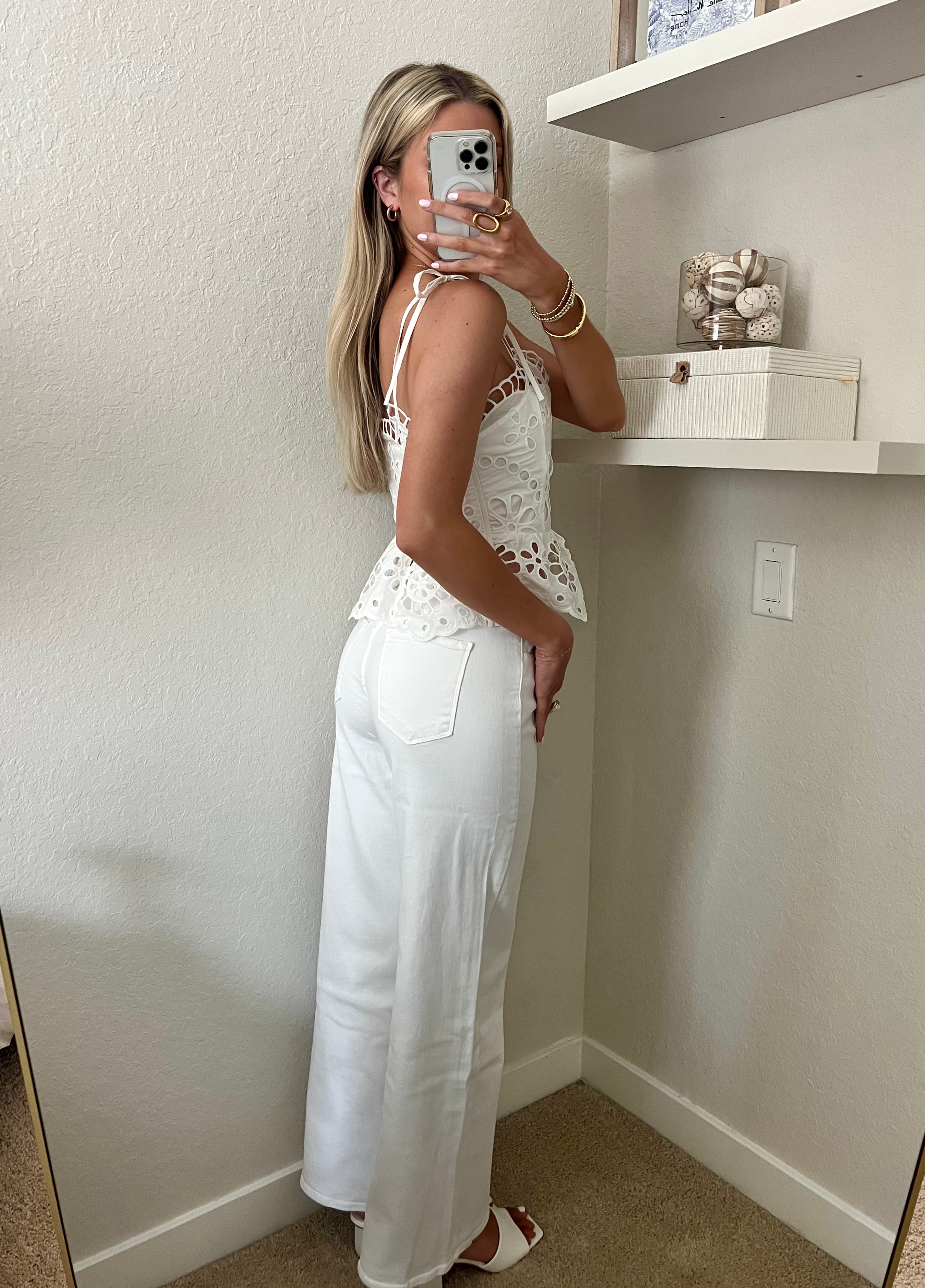 Woman taking a mirror selfie wearing a white lace top and white jeans with heels in a room.