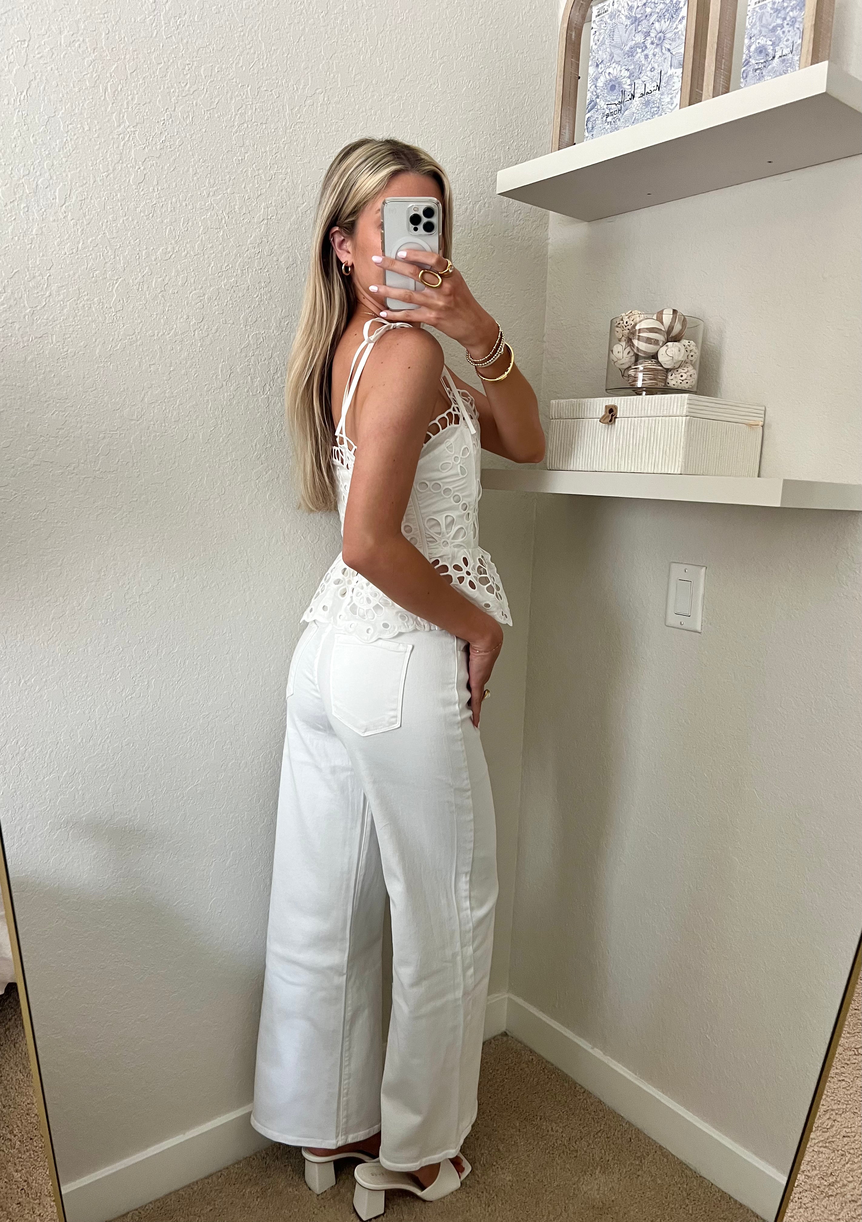 Woman taking a mirror selfie wearing a white lace top and white jeans with heels in a room.