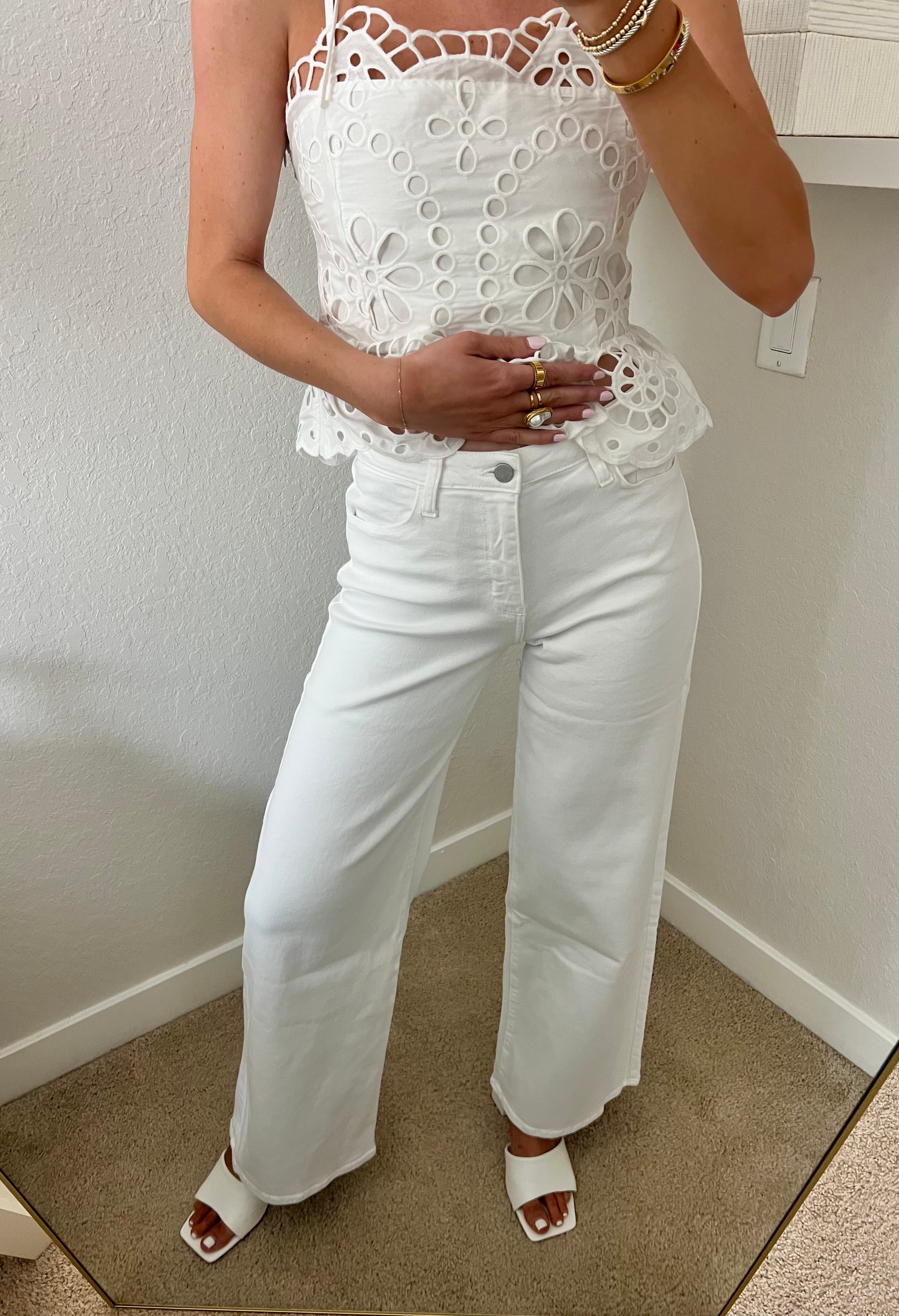 Woman taking a mirror selfie wearing a white lace top and white jeans with heels in a room.