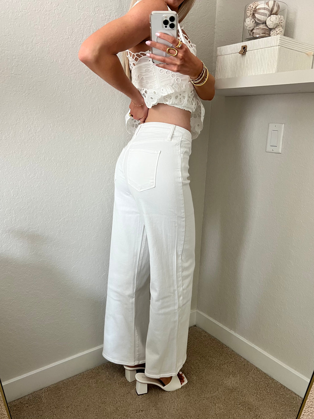 Woman taking a mirror selfie wearing a white lace top and white jeans with heels in a room.