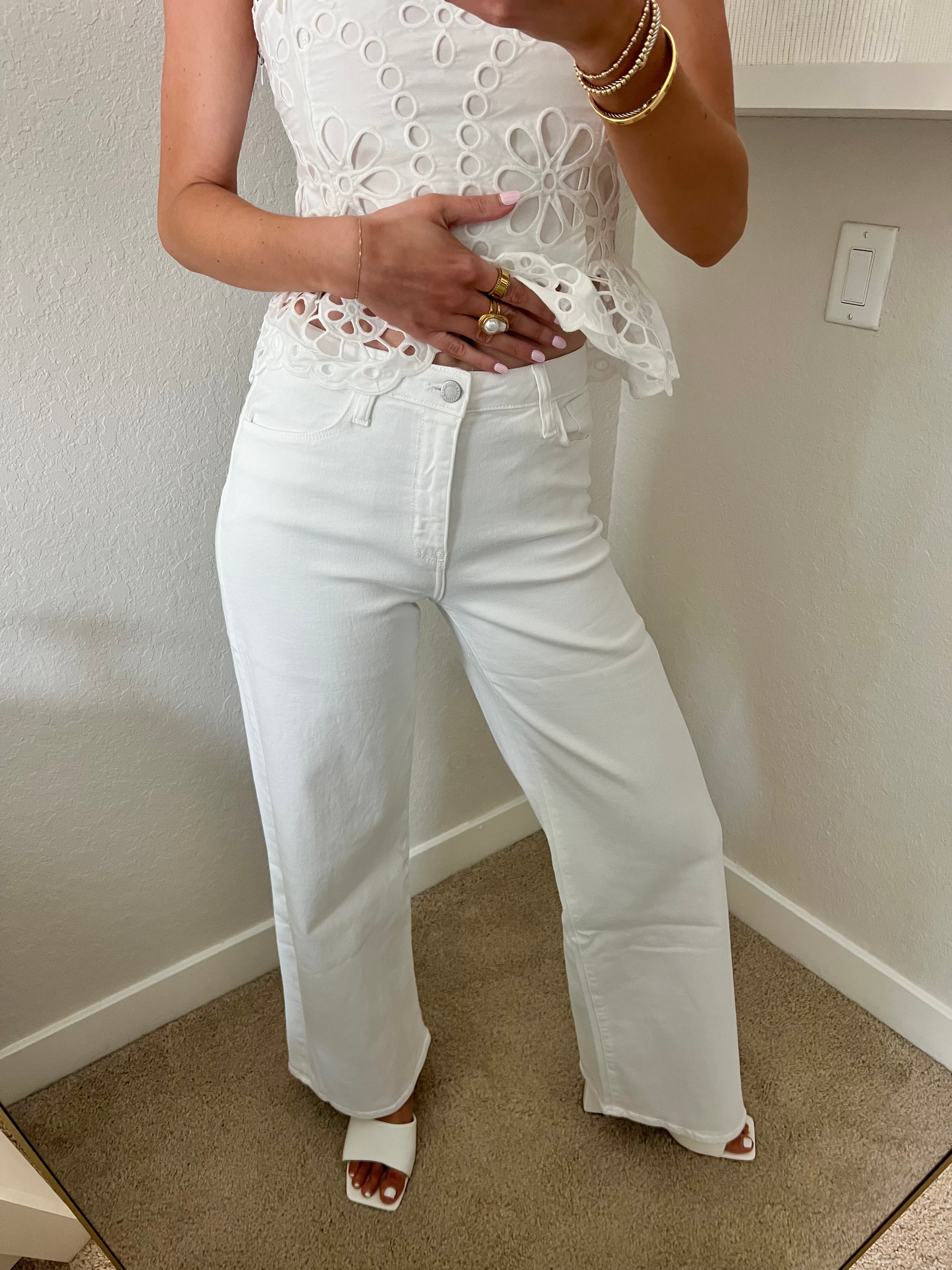 Woman taking a mirror selfie wearing a white lace top and white jeans with heels in a room.
